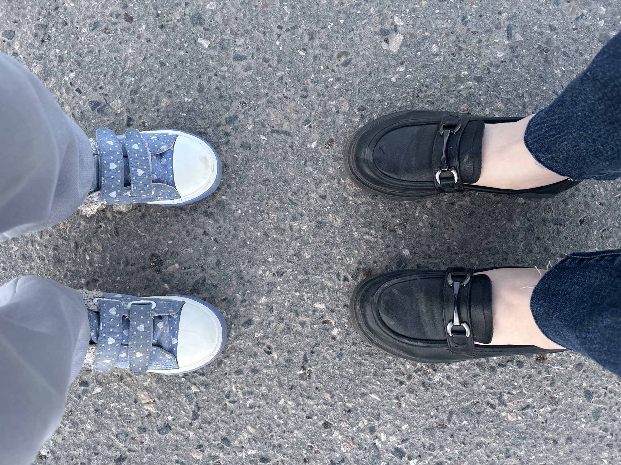 The image shows two pairs of feet standing on an asphalt surface. On the left, a child wears light blue canvas shoes with white hearts and white toe caps, paired with light gray pants. On the right, an adult wears black loafers with a metal buckle detail, paired with blue jeans. The background is a textured asphalt surface with varying shades of gray. The location, Chaikovsky, Russia, cannot be verified from the provided image.