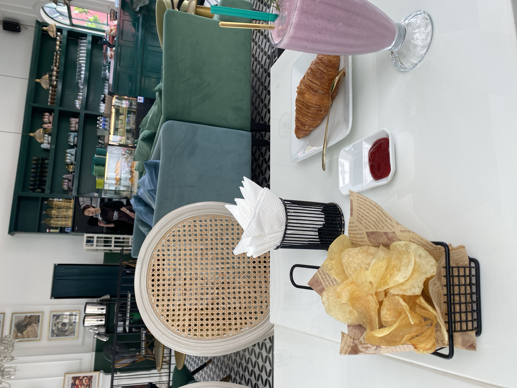 This image captures a bright, modern cafe interior, likely in Da Lat, Vietnam, from a top-down perspective of a white table. The scene is presented as if rotated 90 degrees clockwise.
In the foreground, a white tabletop is laden with various food and drink items. A tall glass contains a purple-pink smoothie with a green straw and a gold spoon. Adjacent to it, a golden-brown croissant rests on a white, irregularly shaped plate, accompanied by another gold spoon. A small, white two-compartment dish holds a dollop of red sauce, likely ketchup or jam. To the right, a black wire basket, lined with brown paper featuring text, is filled with light yellow potato chips. A black wire holder with neatly folded white paper napkins sits beside it. A light-colored rattan-style chair back is partially visible to the left.
The background reveals the cafe's stylish decor. Dark green upholstered chairs and sofas are arranged around tables, contrasting with white walls and a patterned black and white floor. Large dark green shelving units line the walls, displaying an assortment of decorative items including small golden birdcages, metallic containers, and various ceramic pieces. Glass display cases showcase pastries and bottled beverages. In the mid-ground, a person with dark hair, wearing a dark top, stands behind a counter area, appearing to look forward. An arched window allows natural light into the space, indicating it is daytime. Framed artwork or pictures are visible on a wall in the lower left, along with a partial view of a chandelier.
The setup suggests a casual dining experience, perhaps a breakfast, brunch, or snack. Text printed on the paper lining the chip basket is partially visible and includes phrases like "The product to keep fresh," "characteristics," "tender and crisp," "flavor, aroma," "bodied, higher," "base, sugar," "cooling process," and "development," likely describing food qualities.