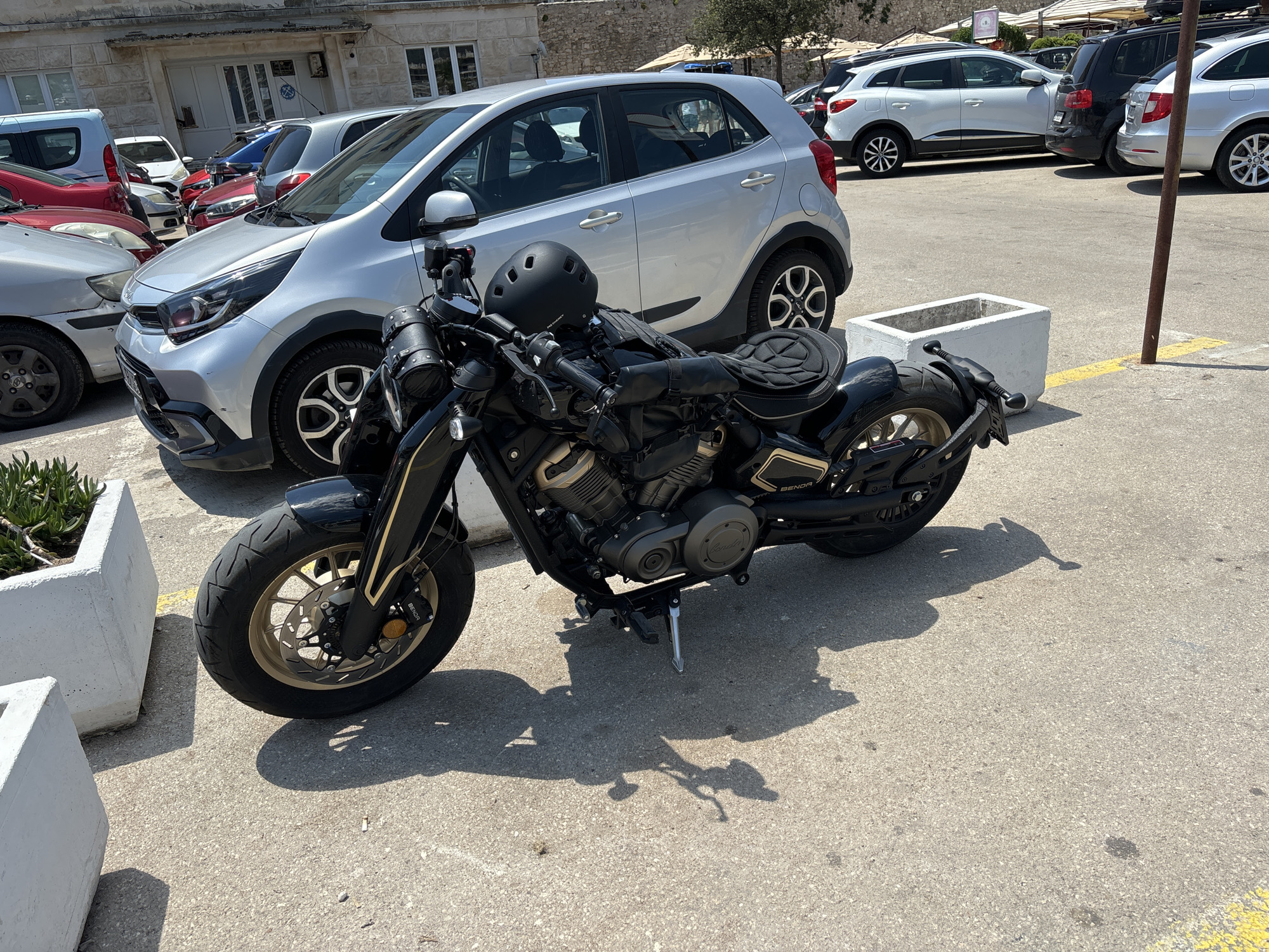 The image depicts a parked motorcycle in a sunny outdoor setting, likely a parking lot. The motorcycle is black with gold accents and appears to be a cruiser-style bike. It has a black helmet resting on its seat.
In the background, several cars are parked, including a silver hatchback, a white SUV, and a dark-colored SUV. The cars suggest a typical urban or tourist area. The architecture in the background, with stone walls and white-framed windows, hints at a Mediterranean or European location.
The scene is illuminated by bright sunlight, casting distinct shadows on the asphalt ground. The weather appears to be clear and warm, typical of a sunny day. The presence of multiple parked vehicles and the general atmosphere suggest this could be a tourist destination, possibly during peak season.
There is no discernible activity taking place; the motorcycle and cars are stationary. The image focuses on the motorcycle in the foreground.
Visible text includes branding on the motorcycle's components, such as "BREMBO" on the brake caliper and "BENDA" on the exhaust pipe. The motorcycle's engine also has a small circular emblem that appears to have text, but it's not clearly legible.