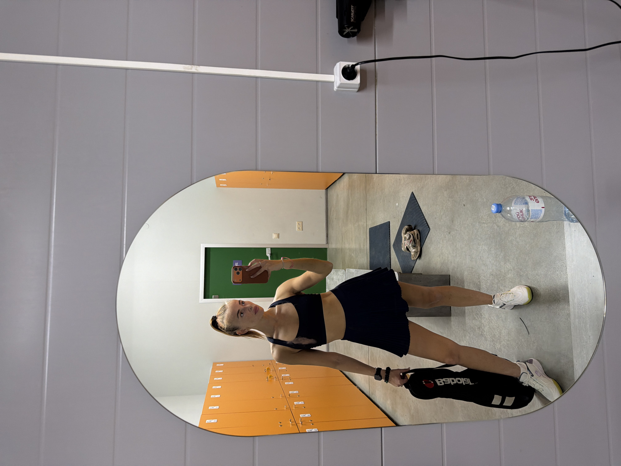 A young woman takes a mirror selfie in a locker room. The walls are gray and there are orange lockers visible in the reflection. She is wearing a black sports bra and skirt, and white sneakers. She holds a black gym bag. A water bottle and a hair dryer are visible on the wall behind the mirror.