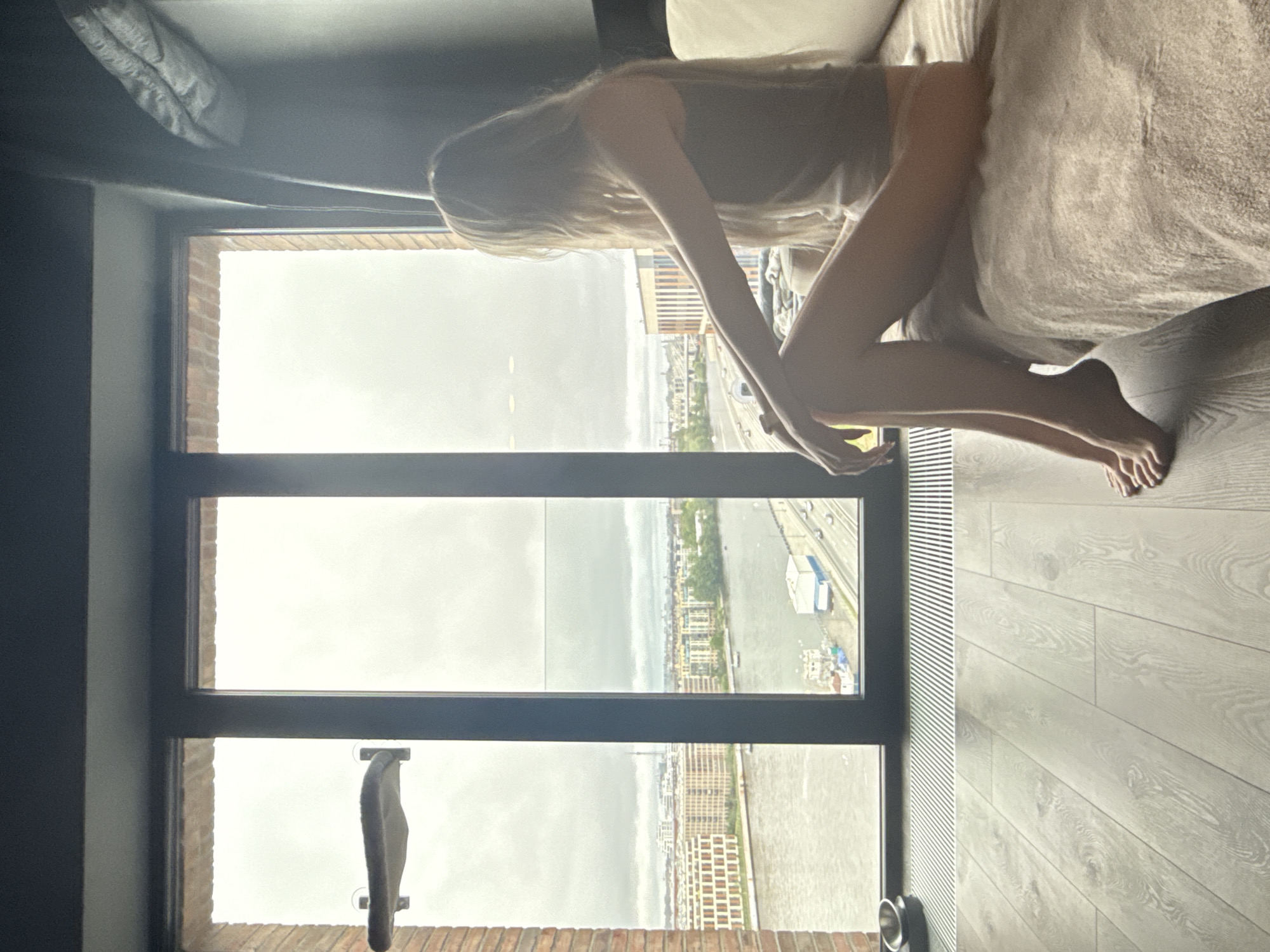 A woman with long blond hair is seated on a bed near a window with a view of St. Petersburg, Russia. She is partially covered by a blanket. The cityscape visible through the window includes a river, buildings, and bridges. The room has a modern interior with wooden floors. There is a radiator and window shades.
