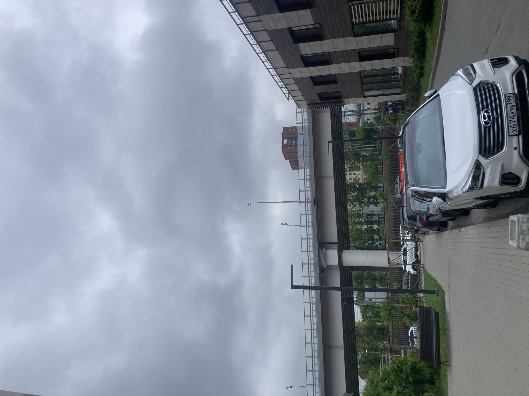 The image shows a street scene in Khimki, Russia on a cloudy day. A white Hyundai car is parked on the right side of the frame, next to a paved sidewalk. Other parked cars are visible behind it. A pedestrian overpass runs above the street, supported by metal pillars. To the left, a modern building with dark gray and light gray rectangular paneling is seen. A bench sits near the sidewalk, amidst green bushes and trees. The sky is overcast with gray clouds. The license plate of the white car is visible and reads "X674YM 799".