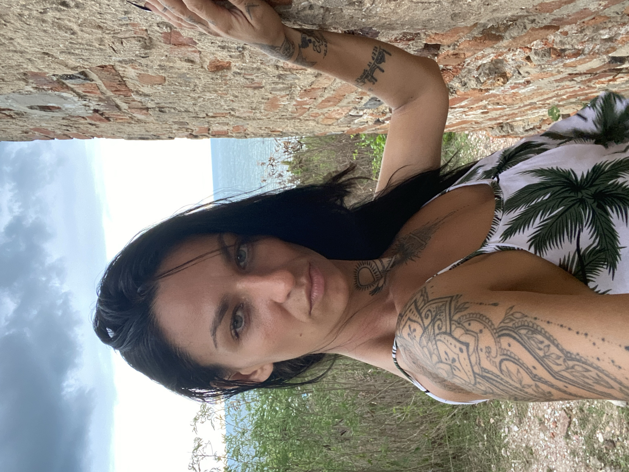 A woman with dark hair and multiple tattoos poses for a photo. She is leaning against a brick wall, with vegetation visible in the foreground and the sea and cloudy sky in the background. She is wearing a white top with a palm tree pattern. The location is Phu Thuy Ward, Vietnam.