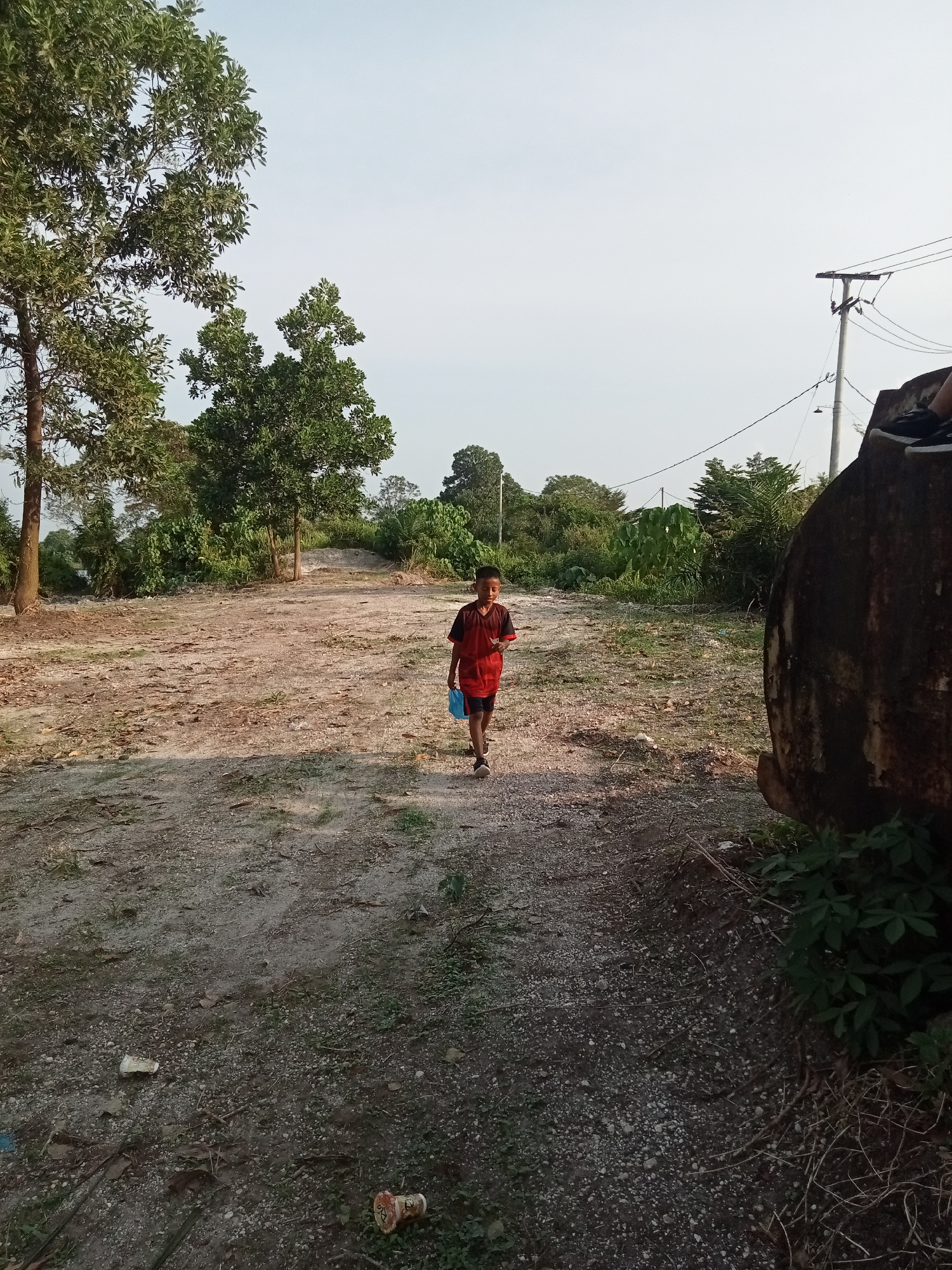 A young boy wearing a red shirt and dark shorts walks on a dirt path in Teluk Kenidai, Indonesia. The boy is carrying a blue plastic bag in his right hand. The path is surrounded by dry vegetation, trees, and some debris. In the background, there are more trees and a utility pole with power lines. To the right of the path, there is a large, dark, round object, possibly a rock or a tank, partially covered by green foliage. The sky is overcast and the lighting suggests it is daytime.