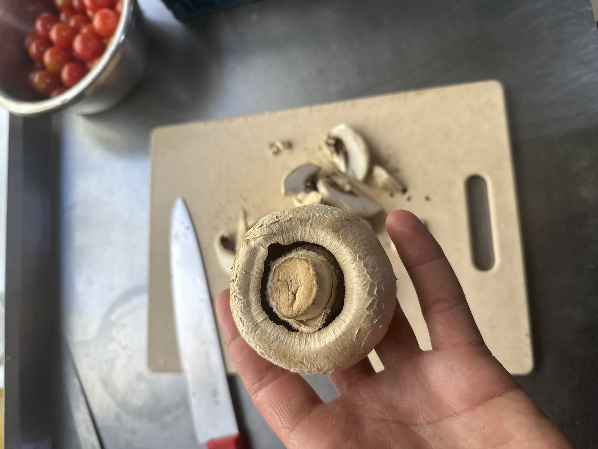 A person's hand holds a single large mushroom with gills visible inside its cap. In the background, sliced mushrooms and a chef's knife lie on a light-colored cutting board. A metal bowl filled with red cherry tomatoes is to the right of the cutting board. The scene appears to be in a kitchen, possibly in Kostanay, Kazakhstan, given the context provided.