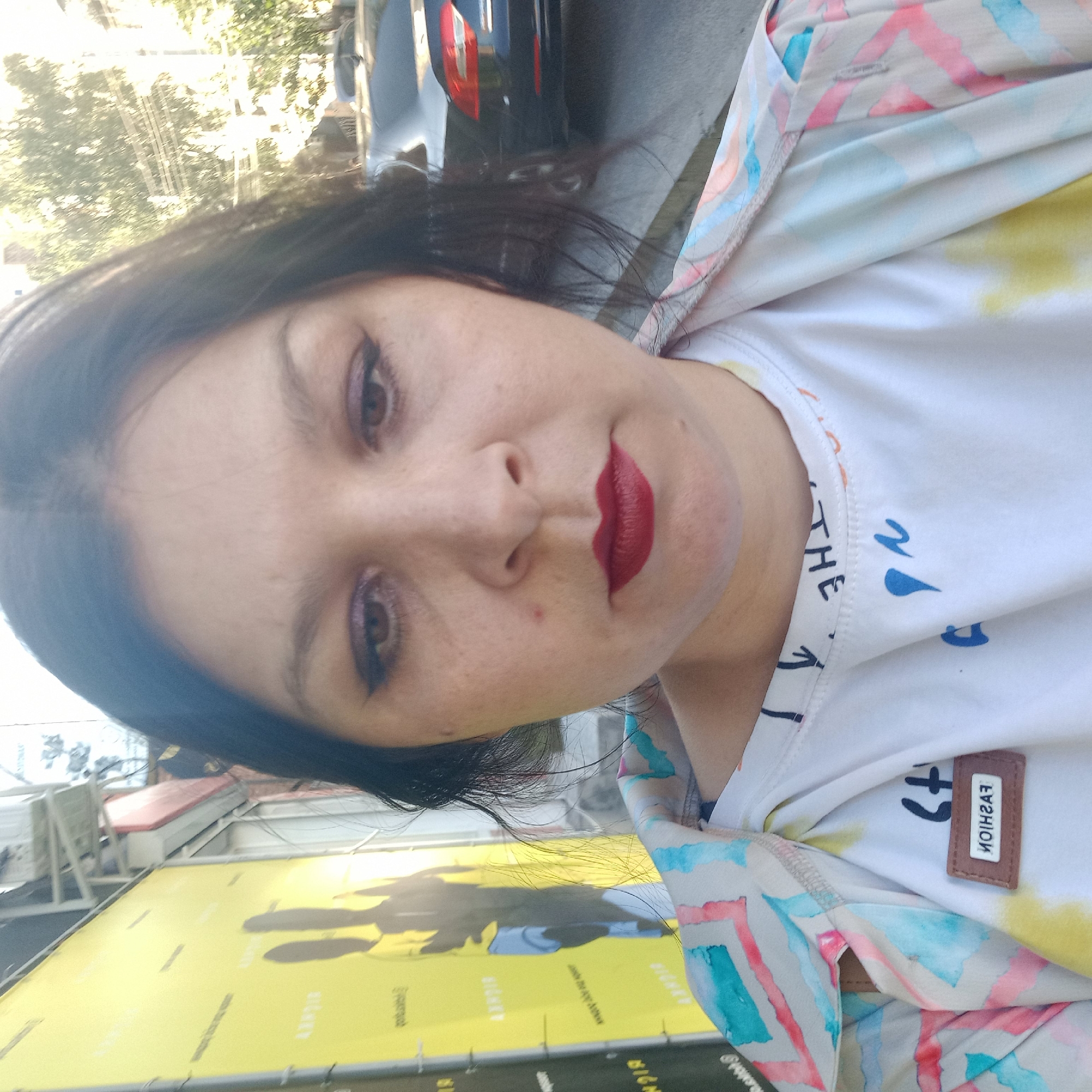 A woman with dark hair and dark brown eyes is taking a selfie outdoors. She is wearing makeup, including eyeshadow and lipstick. She is wearing a white t-shirt with colorful abstract patterns, and a matching patterned jacket is draped over her shoulders. In the background, there is a yellow billboard with silhouettes of people, a black car, trees, and buildings. The location is specified as Kropyvnytskyi, Ukraine.