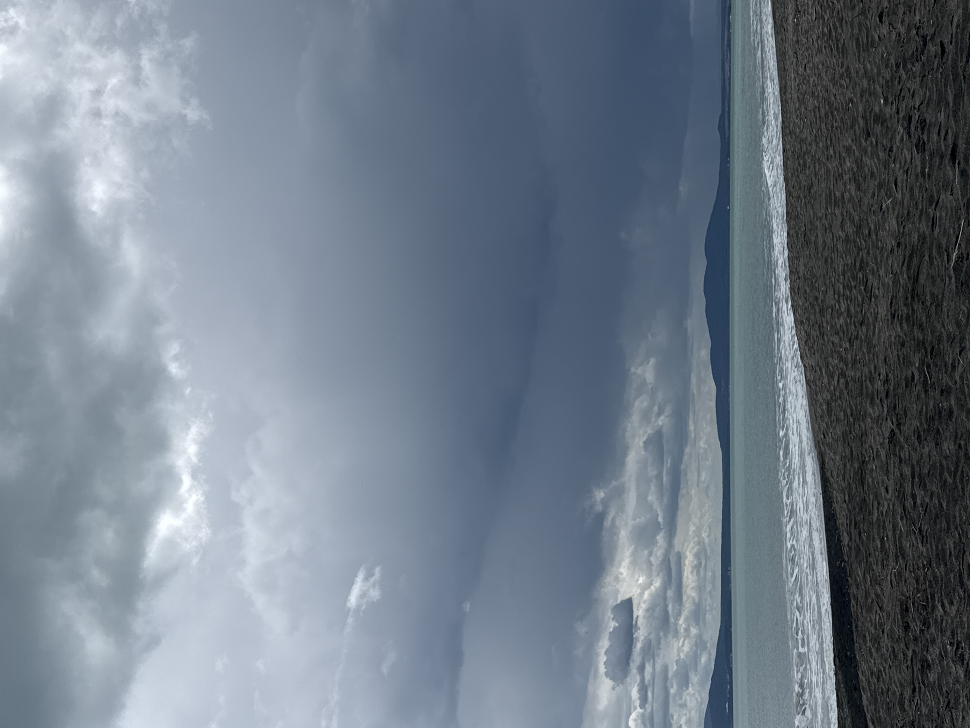 The image depicts a coastal scene. A dark, granular beach occupies the right side. A light-colored shoreline and ocean are visible to the left of the beach. Further to the left, a strip of land and hills are visible. The sky dominates the left portion of the image, filled with a mix of dark, heavy storm clouds and patches of lighter, brighter clouds.
Based on the available information, a definitive location in Burgas, Bulgaria, cannot be determined. Burgas is a coastal city with beaches and open sea views, which are consistent with the image. Further contextual information would be needed to determine the exact location.