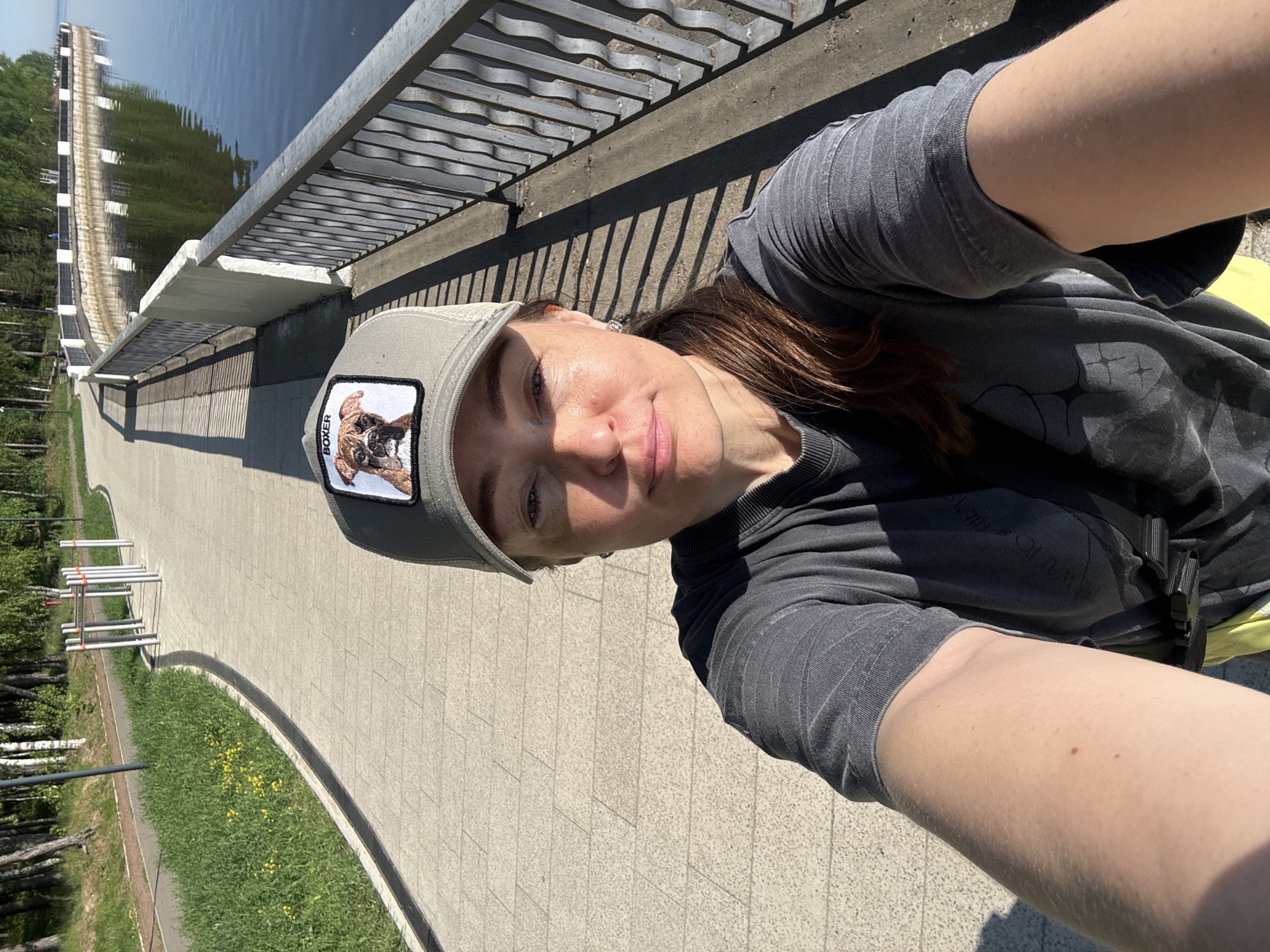 A woman is taking a selfie outdoors, likely in the city of Chaikovsky, Russia. She is wearing a gray t-shirt, a cap with a dog patch, and is positioned near a railing, sidewalk, grass, and trees. In the background, a body of water and what appears to be a dam structure are visible. The photo is rotated, with the ground appearing vertical.