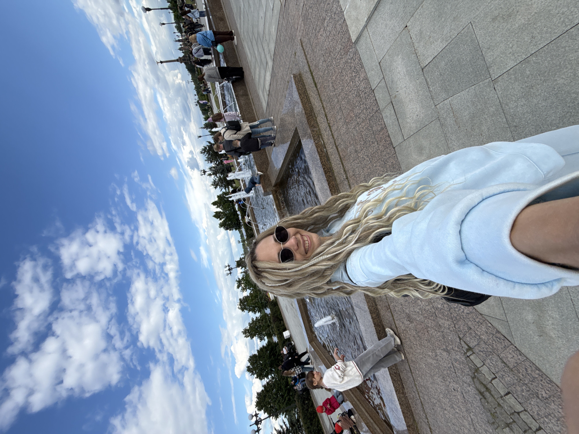 The image captures a sunny day at a public fountain in Rusanovo, Russia. The focal point is a woman with blonde dreadlocks and sunglasses, presumably taking a selfie. Behind her, the fountain features multiple tiers of water jets. A group of people, including adults and children, are gathered near the fountain, some interacting with the water. The background includes trees, lampposts, and other structures consistent with a city park or plaza. The sky is blue with scattered clouds. The pavement surrounding the fountain is made of granite tiles.