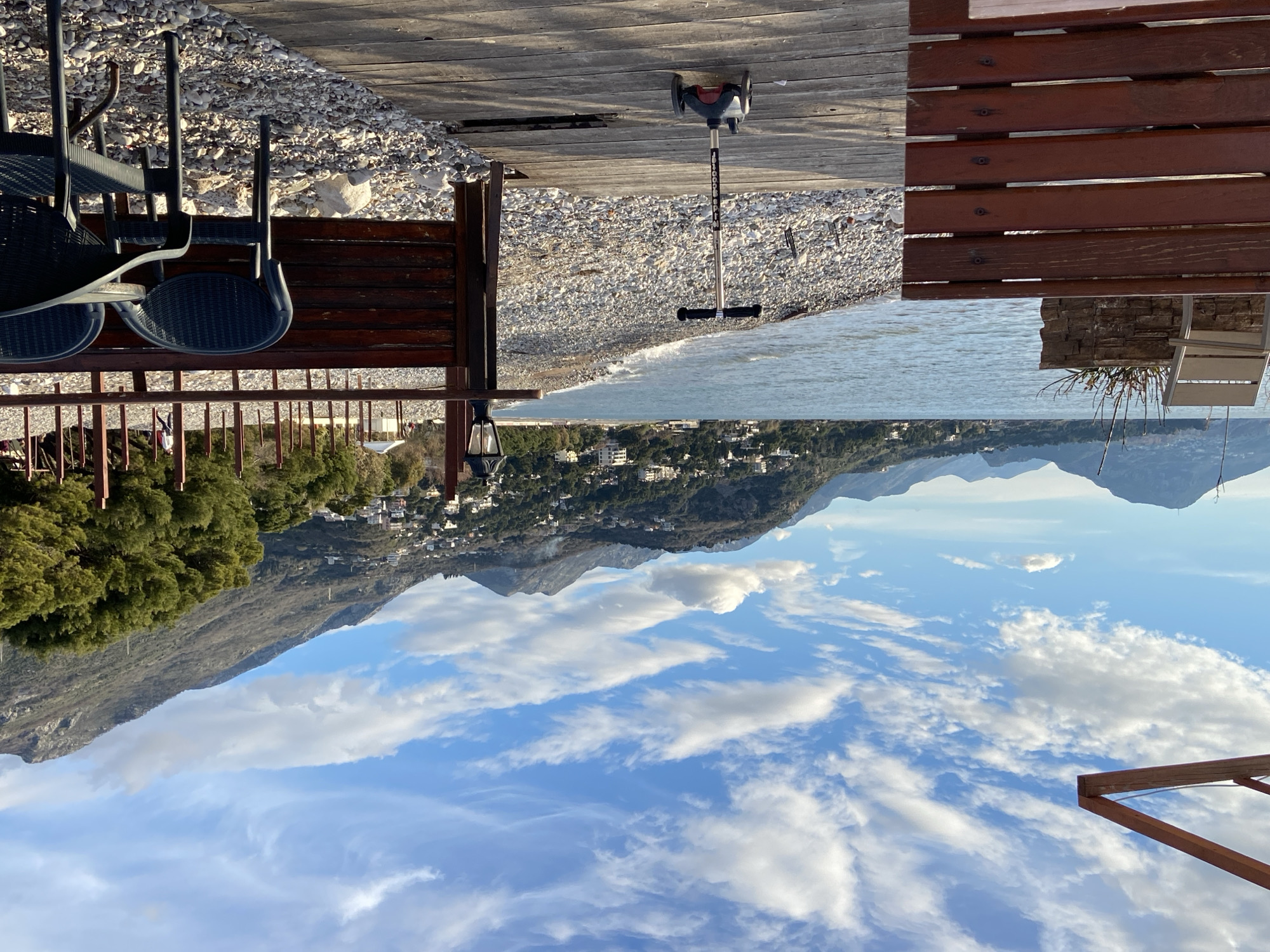 The image shows a topsy-turvy view of a scene likely in the coastal city of Bar, Montenegro. In the original orientation, the sky is at the bottom with scattered white clouds against a blue backdrop. Above the sky, a mountain range extends across the scene, dotted with small buildings that suggest a populated area on the slopes. The image is bisected by a reversed shoreline, with a pebble beach transitioning into the ocean. At the top of the image, upside-down tables, chairs, and wooden planks create a foreground. A folded scooter rests on the wooden planks. The overall scene suggests a coastal restaurant or terrace with a scenic view.