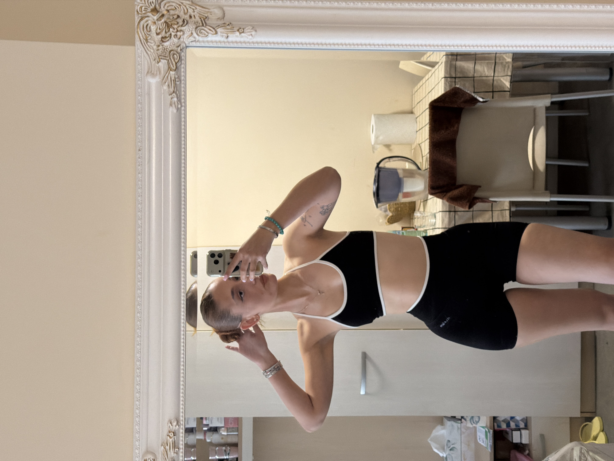 A young woman stands in what appears to be a bathroom, taking a mirror selfie. She is dressed in a black sports bra with white trim and black shorts. The mirror is ornate and framed in white. The background includes a toilet with a white seat and a brown towel, a roll of toilet paper, a vacuum cleaner, and various bathroom cabinets and items on the shelves below. The location appears to be inside a domestic residence, but there is no information available to suggest that it is in the city of Sofia, Bulgaria.