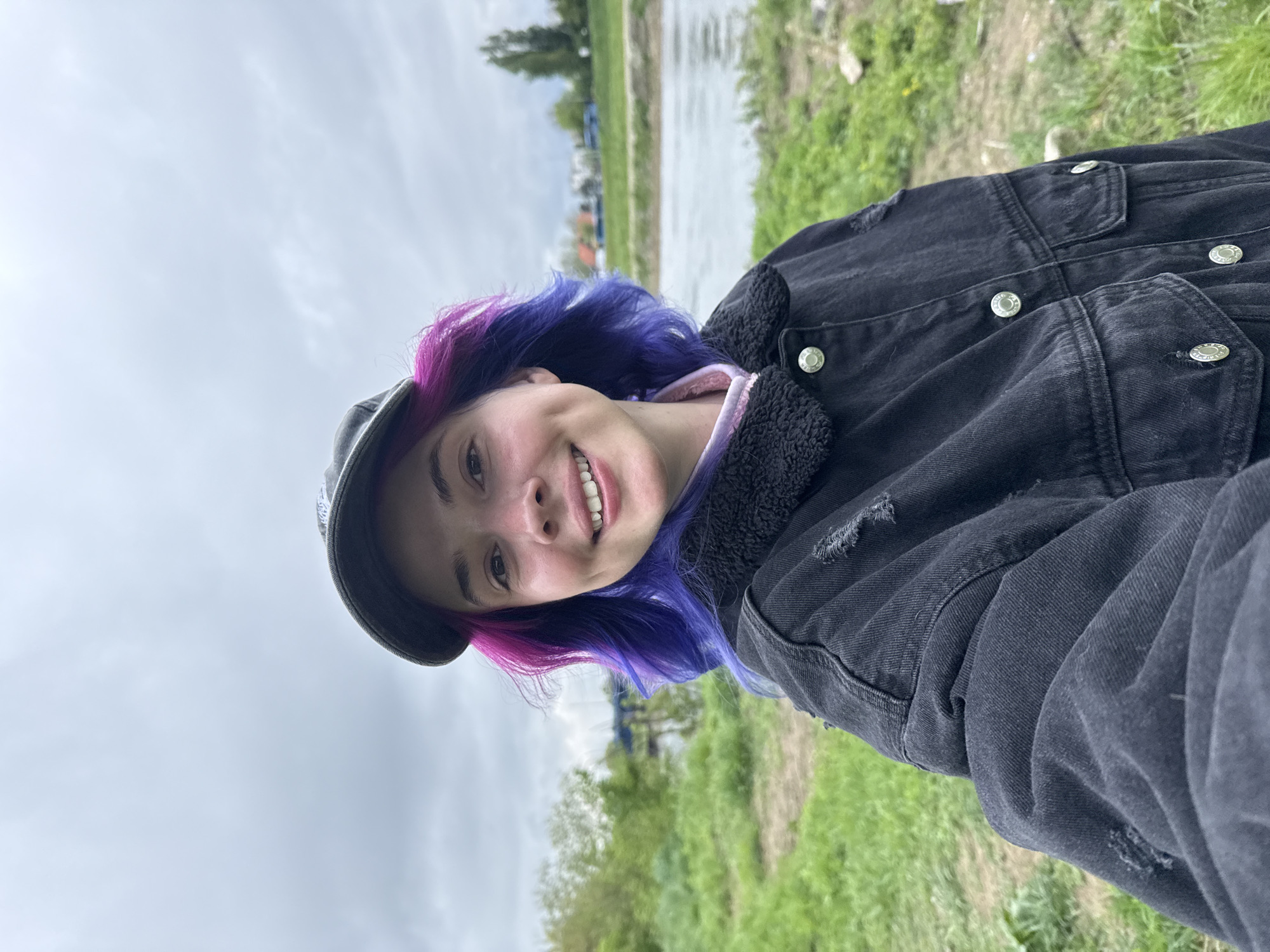 A close-up shot, rotated sideways, depicts a smiling person outdoors. They have short hair colored in a blend of purple and blue. They are wearing a black denim jacket with silver buttons and a black cap.
The background suggests an outdoor environment. The sky is overcast. A body of water is visible, as is green grass and some trees.
The image was taken in the City of Zagreb, Croatia.