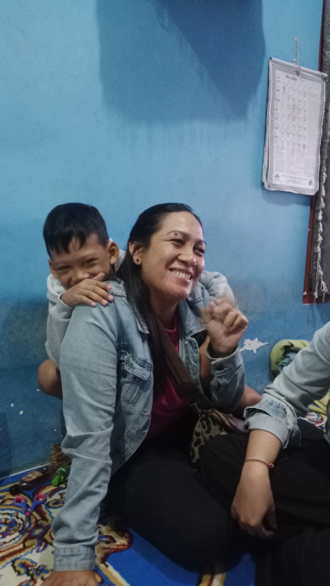 A medium shot captures a boy on a woman's back, both facing forward, against a blue wall in what appears to be a room in Denpasar, Indonesia. The woman, positioned slightly to the right, wears a denim jacket over a pink shirt, with her head tilted up and a wide smile. She has dark hair pulled back and visible earrings. Her right hand is raised, with fingers extended, as if waving or gesturing. The boy, on her left, peeks over her shoulder, his head tilted and a playful grin on his face. He is wearing a light-colored hoodie. A brown strap from a bag is visible across the woman's chest, and a patterned bag rests between her and the boy. To the right, a portion of another person's arm, wearing a similar denim jacket, is visible, along with dark pants. On the wall behind them, a calendar for July 2026 is hanging, and a colorful rug is visible on the floor. Several items, including a green bottle, a pair of scissors, and a bottle of liquid, are scattered on the floor near the rug. The overall impression is a candid moment of joy and connection within a domestic setting.