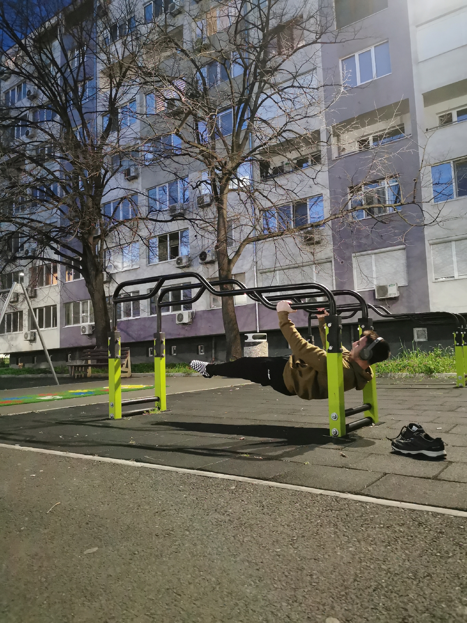 A person wearing a brown jacket, black pants, and headphones is hanging horizontally from a black and green metal outdoor exercise structure. Their legs are straight and extended, feet pointed to the left. The ground appears to be a dark rubberized material. A pair of black and white sneakers are lying on the ground next to the structure's base. In the background is a large apartment building with light-colored walls and numerous windows, as well as leafless trees. Based on the building style and layout, the location is likely in a residential area of Burgas, Bulgaria. The lighting suggests it is either dusk or dawn.