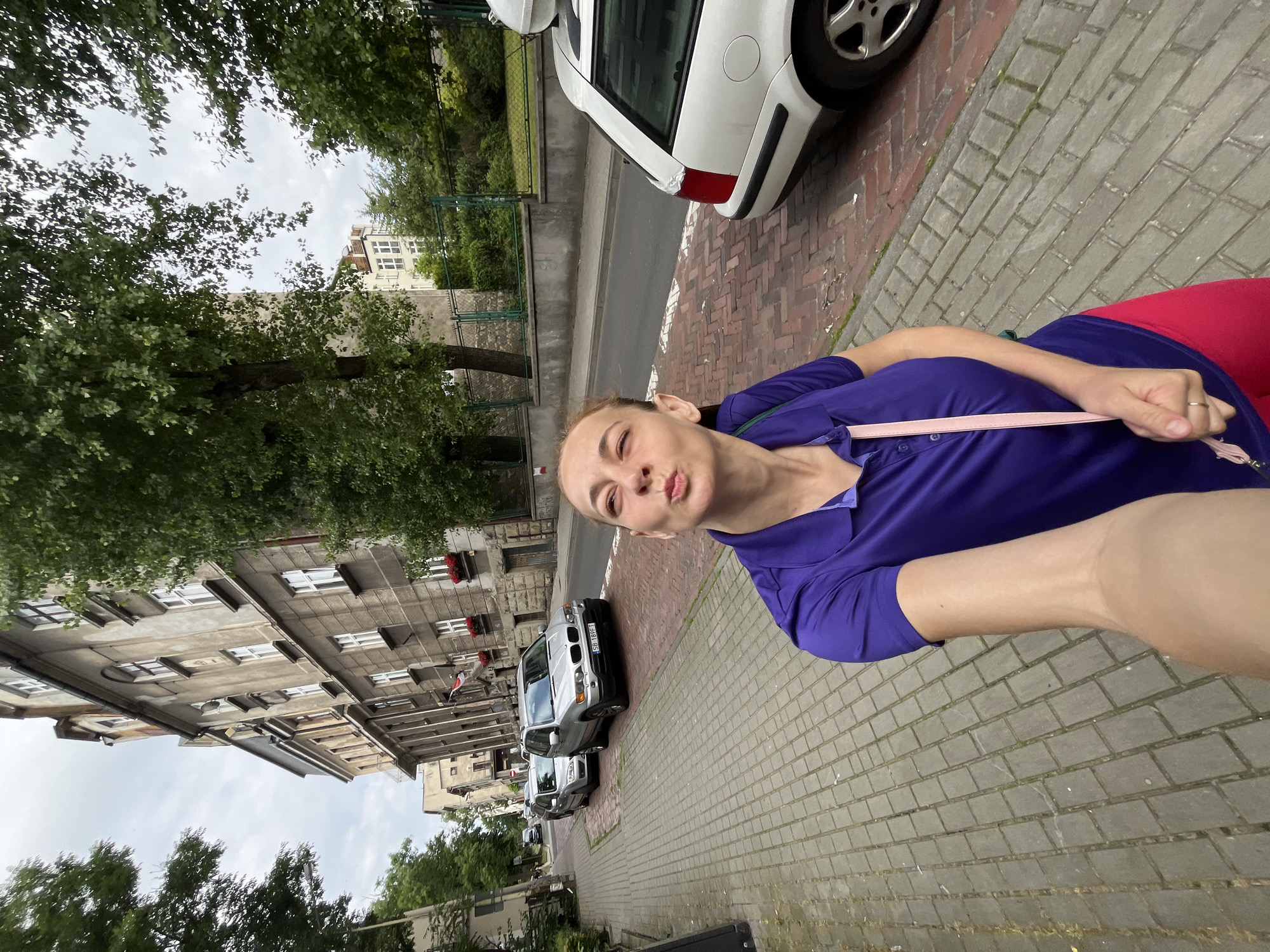 A woman is taking a selfie with her mouth puckered in a kiss pose while standing on a cobblestone sidewalk. She is wearing a purple polo shirt and bright pink leggings, with a light pink strap across her chest. In the background, there is a street with parked cars, including a silver SUV and a white station wagon. Buildings line the street, some with visible windows and red flower boxes. Trees with green foliage are present on both sides of the street, and the sky is overcast.