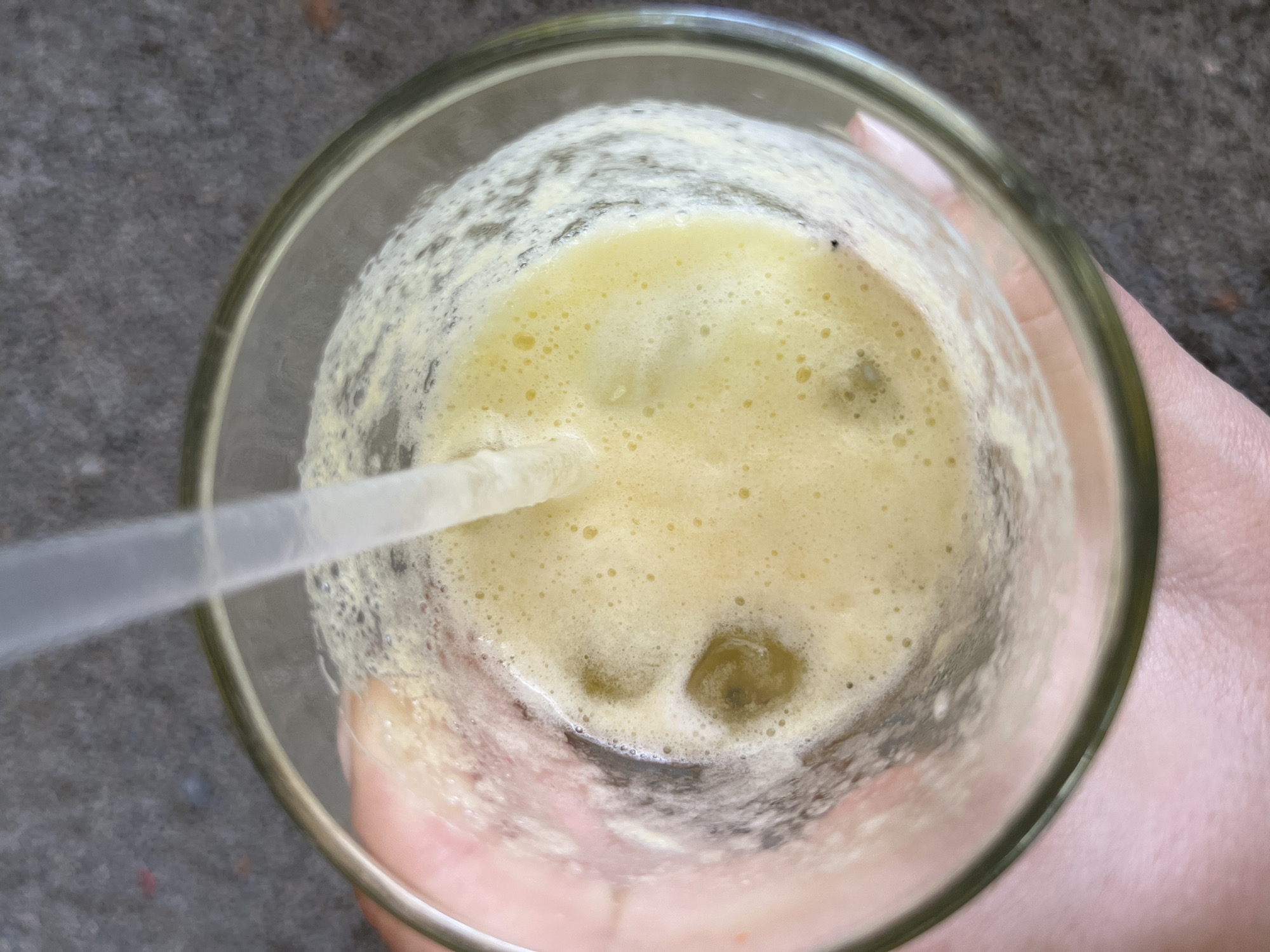 A close-up, overhead shot shows a glass of yellowish-green liquid with a straw in it, held by a person. The liquid is foamy, and there are some dark green or brown solids settled at the bottom of the glass. The glass itself is clear, and the straw is translucent with a slight gradient. The person's hand is visible holding the glass; the skin tone appears fair. The background consists of a dark gray surface.