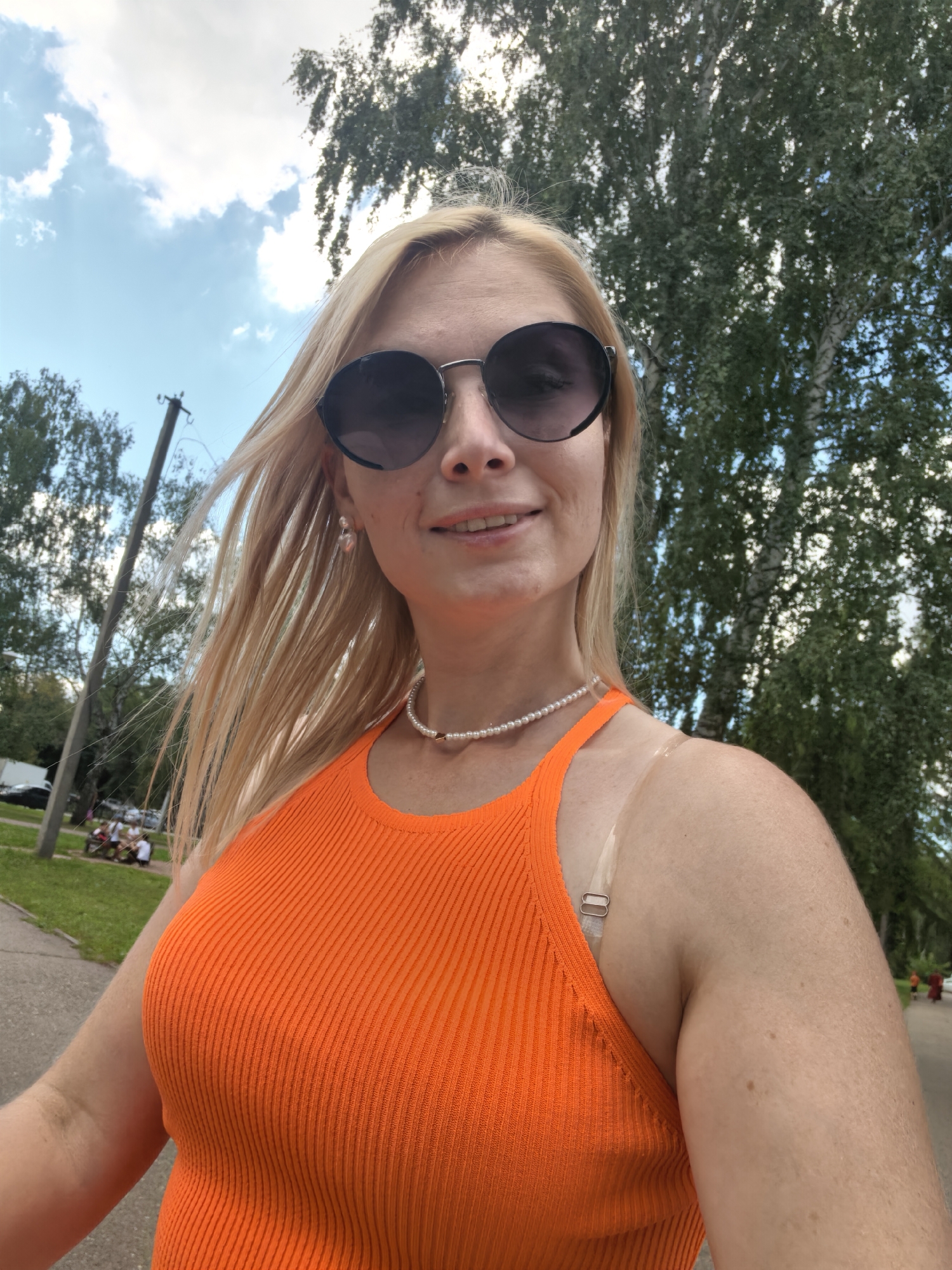 A young woman with blonde hair, wearing sunglasses, an orange ribbed tank top, and a pearl necklace, smiles at the camera. Her left shoulder shows a tan line. She is outdoors, with trees and a cloudy sky in the background. A utility pole is visible to her left. In the background, there's a grassy area with people sitting on benches, and a paved path. The setting appears to be a park or recreational area.