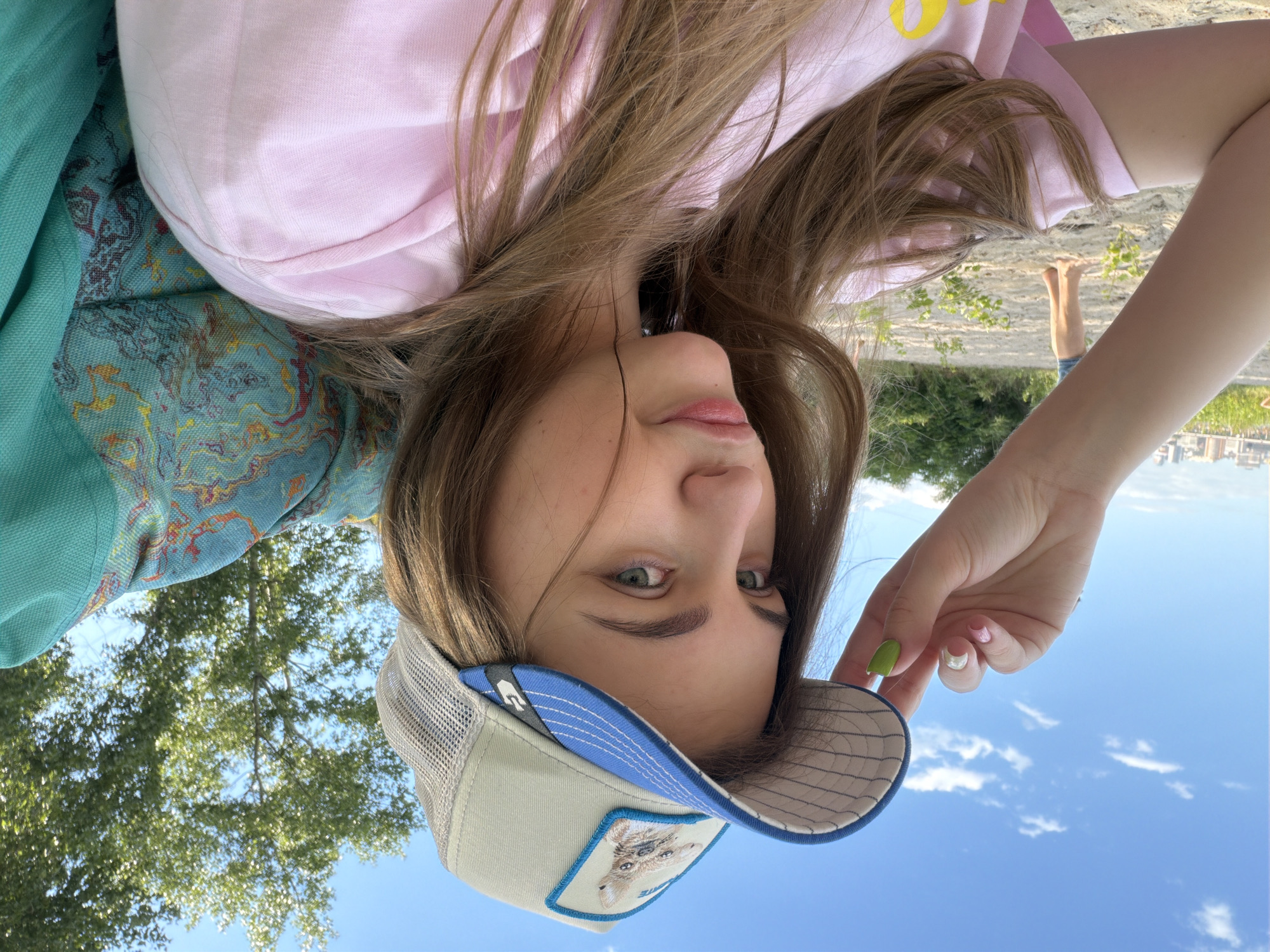 A young woman with long, brown hair and light-colored eyes is taking a selfie outdoors. She is wearing a light pink t-shirt and a beige trucker hat with a picture of a dog on it. The hat has a blue brim and a grey mesh back. She is holding the brim of the hat with her left hand, which has painted nails in pink, white, and green. She is looking towards the camera with a slightly pensive expression. Behind her, on the left, is a sandy area with some sparse green vegetation and a distant view of buildings, suggesting an urban setting. Further back, a person's legs are visible, standing on the sand. To the right of the woman is a patterned teal fabric, possibly part of a chair or blanket. The background is a bright blue sky with scattered white clouds and the lush green foliage of a tree. This scene is consistent with a location along the Dnipro River embankment in Dnipro, Ukraine, which often features sandy beaches and views of the city skyline.