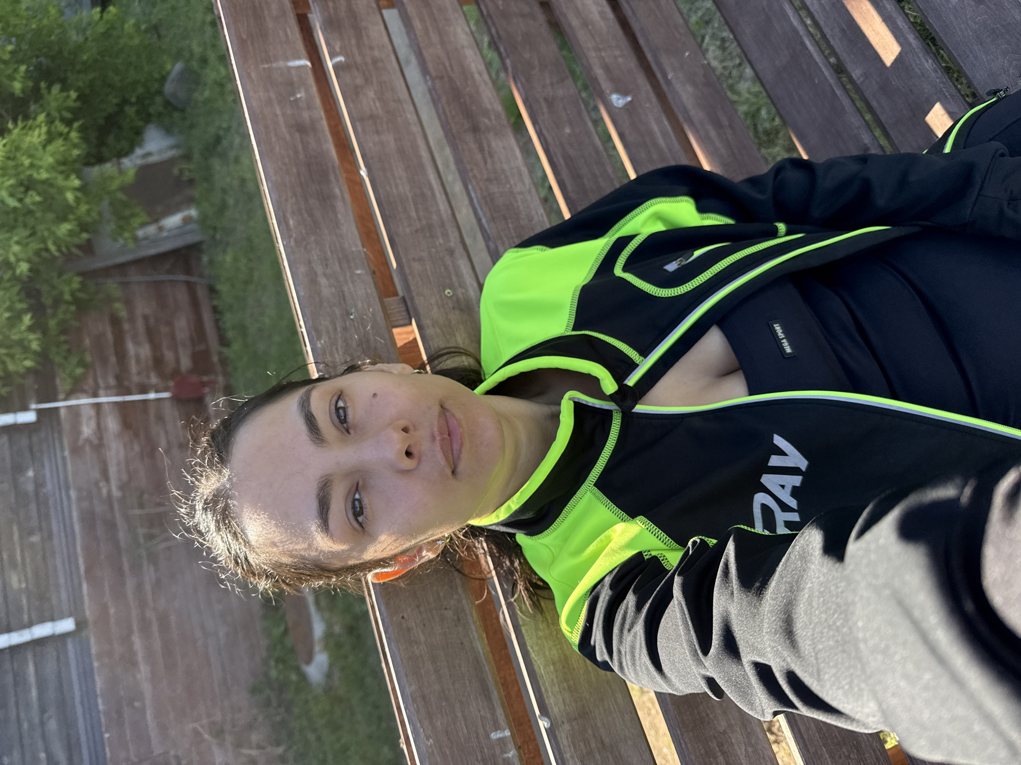 A woman is sitting on a wooden bench outdoors, on a sunny day. She is wearing a black and neon green athletic jacket over a black sports bra. The jacket has a zipper and a logo that reads "RAY" in white letters. She is looking directly at the camera with her hair pulled back. The background consists of a wooden fence and greenery. The image appears to have been taken in Chaikovsky, Russia, given the context provided.