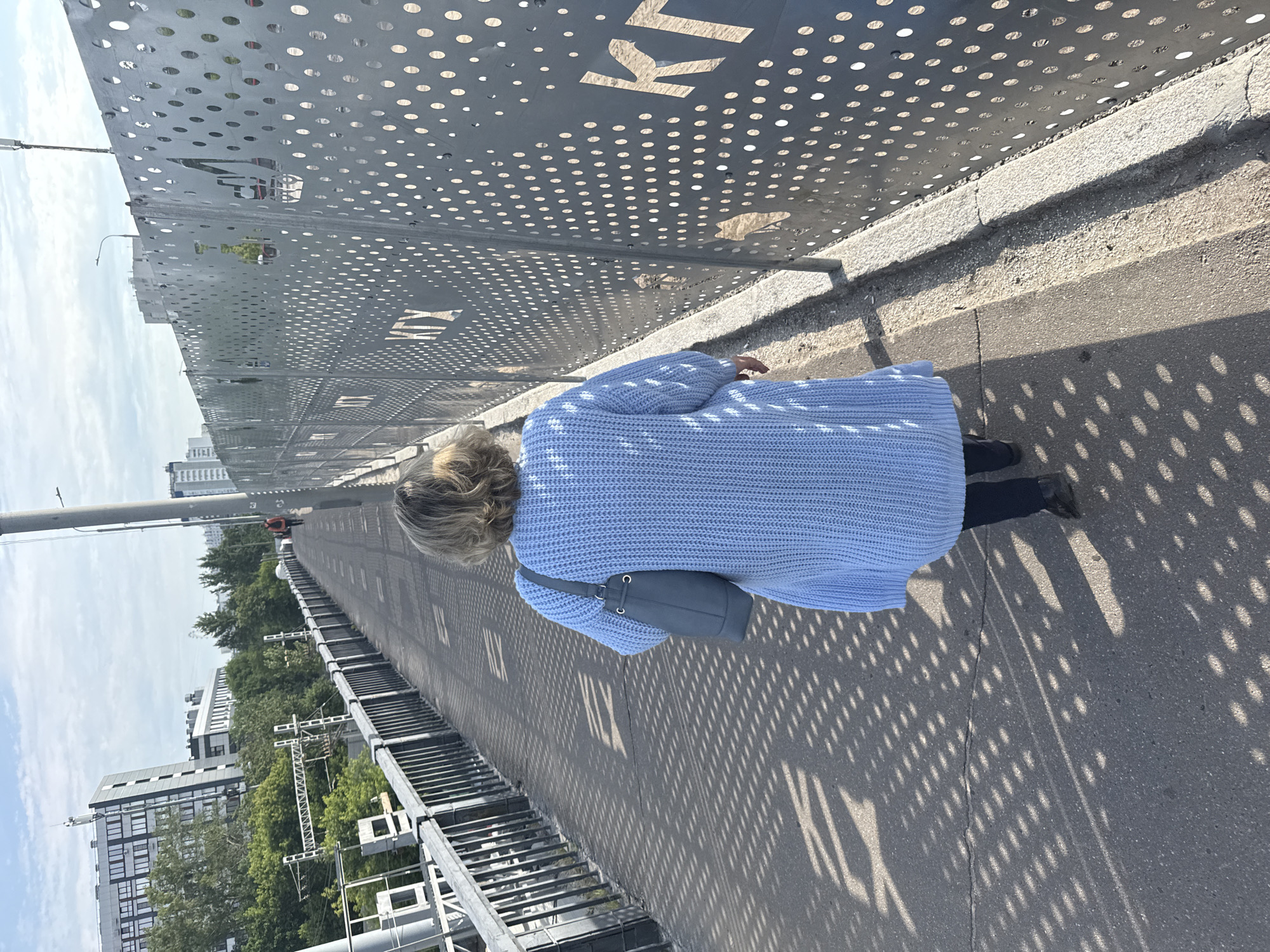 A middle-aged woman walks away from the camera on a pedestrian bridge in Moscow, Russia. She is wearing a light blue knitted cardigan and dark pants, carrying a gray purse. The bridge has a metal railing on the left and a decorative metal fence with circular perforations on the right, on which the letters "KTX" are visible. The ground of the bridge is asphalt with a pattern of shadows cast by the fence. In the background, there are buildings, trees, and railway tracks, indicating an urban environment. The sky is cloudy.