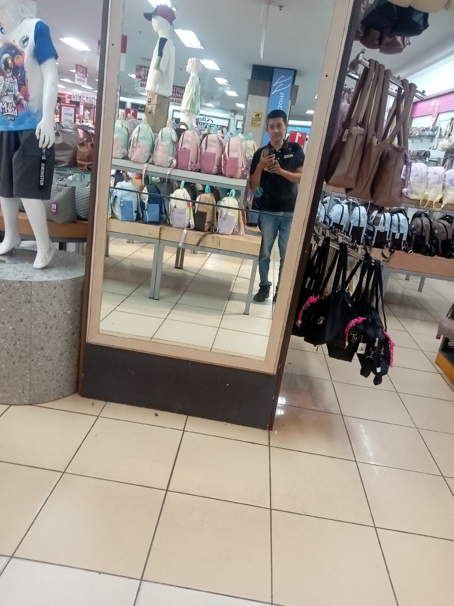 The image depicts a man taking a selfie in a retail store that sells bags and clothing. The store appears to be located in Batam, Indonesia, based on the Indonesian text visible in the background, such as "MURAH BANGED" (very cheap) and "DISKON" (discount). In the foreground, a mannequin dressed in a t-shirt with an astronaut graphic and shorts is visible. The main subject, the man, is standing in front of a large mirror, wearing a black polo shirt and blue jeans, and holding a smartphone to take the picture. He is reflected in the mirror along with displays of bags and backpacks. The store environment includes tiled flooring and various shelves and racks displaying merchandise.