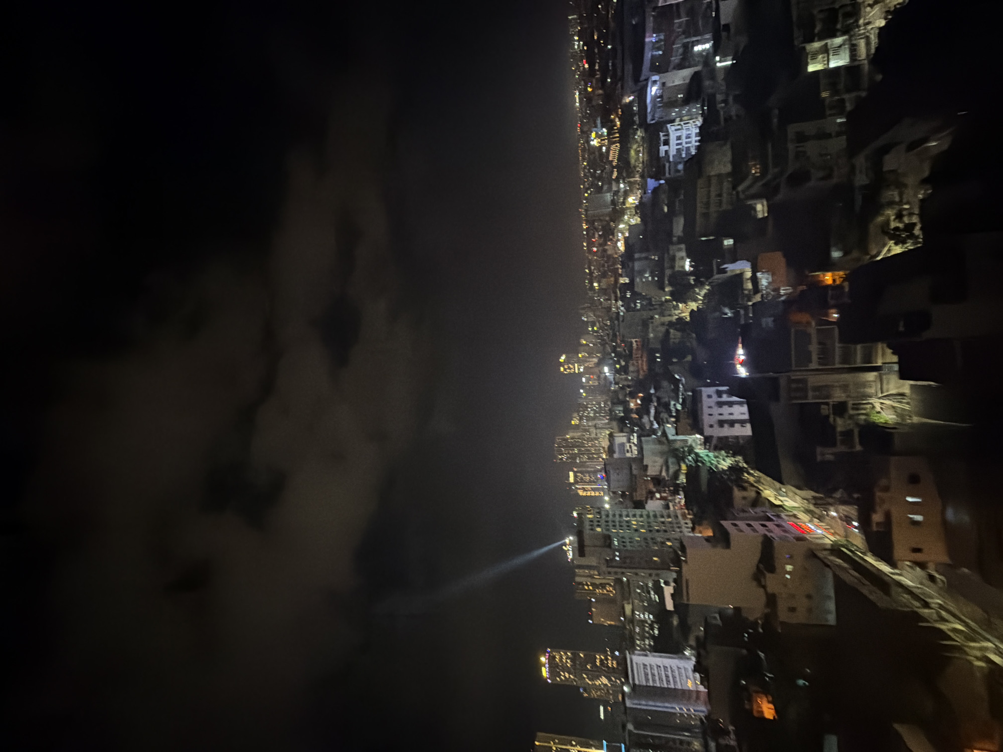 The image displays an elevated, nighttime view of an urban area within Nha Trang Ward, Vietnam. The upper portion of the frame is dominated by a dark sky with visible, textured cloud cover. A singular, intense beam of white light emanates vertically from the pinnacle of a multi-story building situated in the mid-left section of the image, extending upwards into the clouds.
The cityscape below encompasses a dense arrangement of structures. In the distant background and mid-ground, numerous high-rise buildings and skyscrapers are illuminated with warm, golden, and white lights, forming a widespread urban horizon. Closer to the foreground, a network of lower to mid-rise buildings is visible, exhibiting a mix of lit and unlit windows and facades. Linear patterns of streetlights and other artificial illumination delineate presumed roadways. No individuals, specific vehicles in motion, or explicit actions are discernible within the frame.