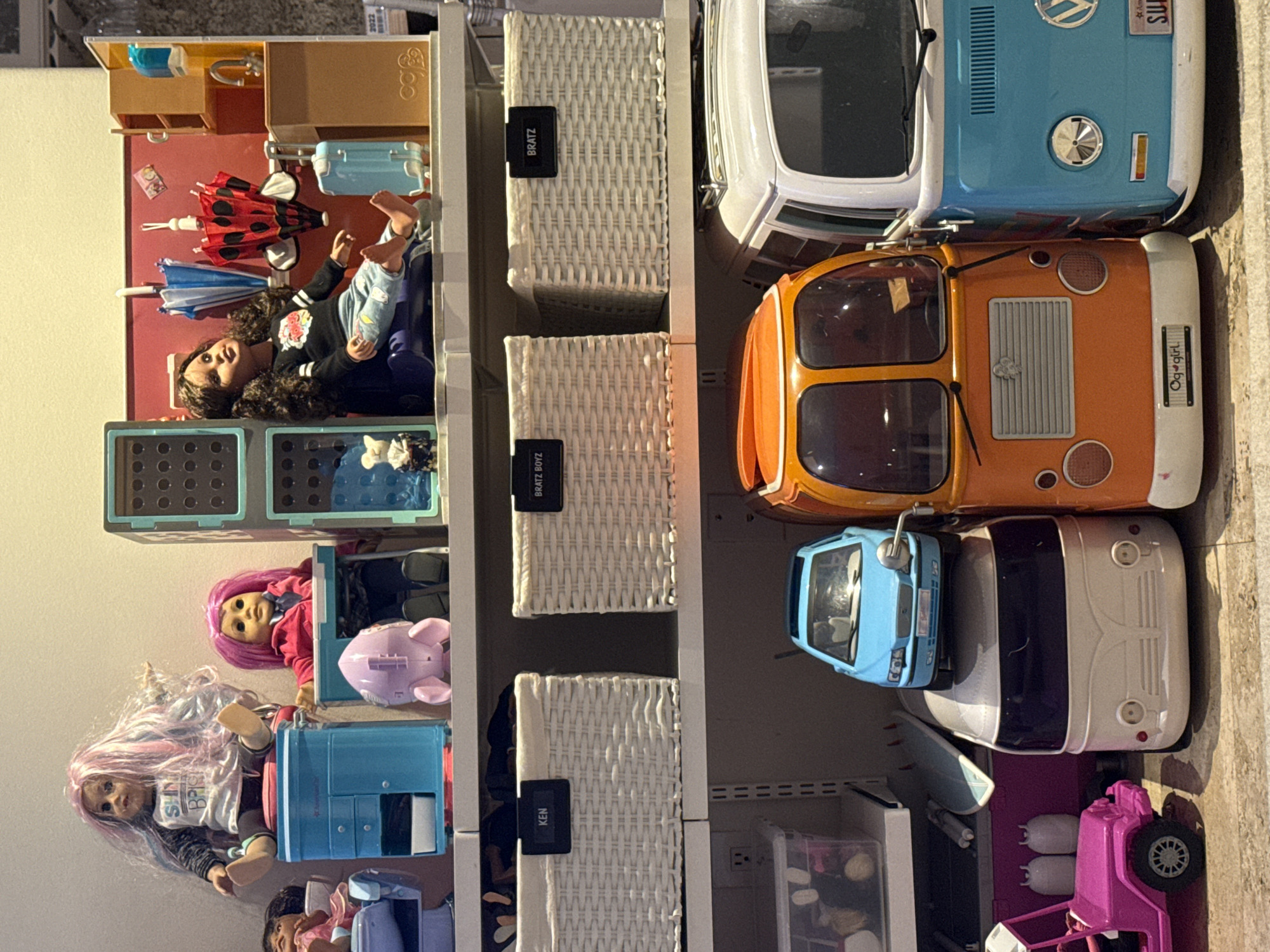 The image depicts a shelf unit against a neutral-colored wall, displaying an assortment of toys and storage containers.
On the left side of the shelf are various dolls and doll accessories. There's a dollhouse-like structure at the top, followed by smaller doll furniture pieces and dolls with different hair colors (purple, pink).
To the right of the doll display are three white woven storage baskets, labeled "BRATZ", "BRATZ BOYZ", and "KEN".
On the far right, there are several toy vehicles. These include a blue and white Volkswagen van, an orange van, a blue car, a white van, and a pink toy car.
There is no indication of the location being Miami Beach, United States of America.