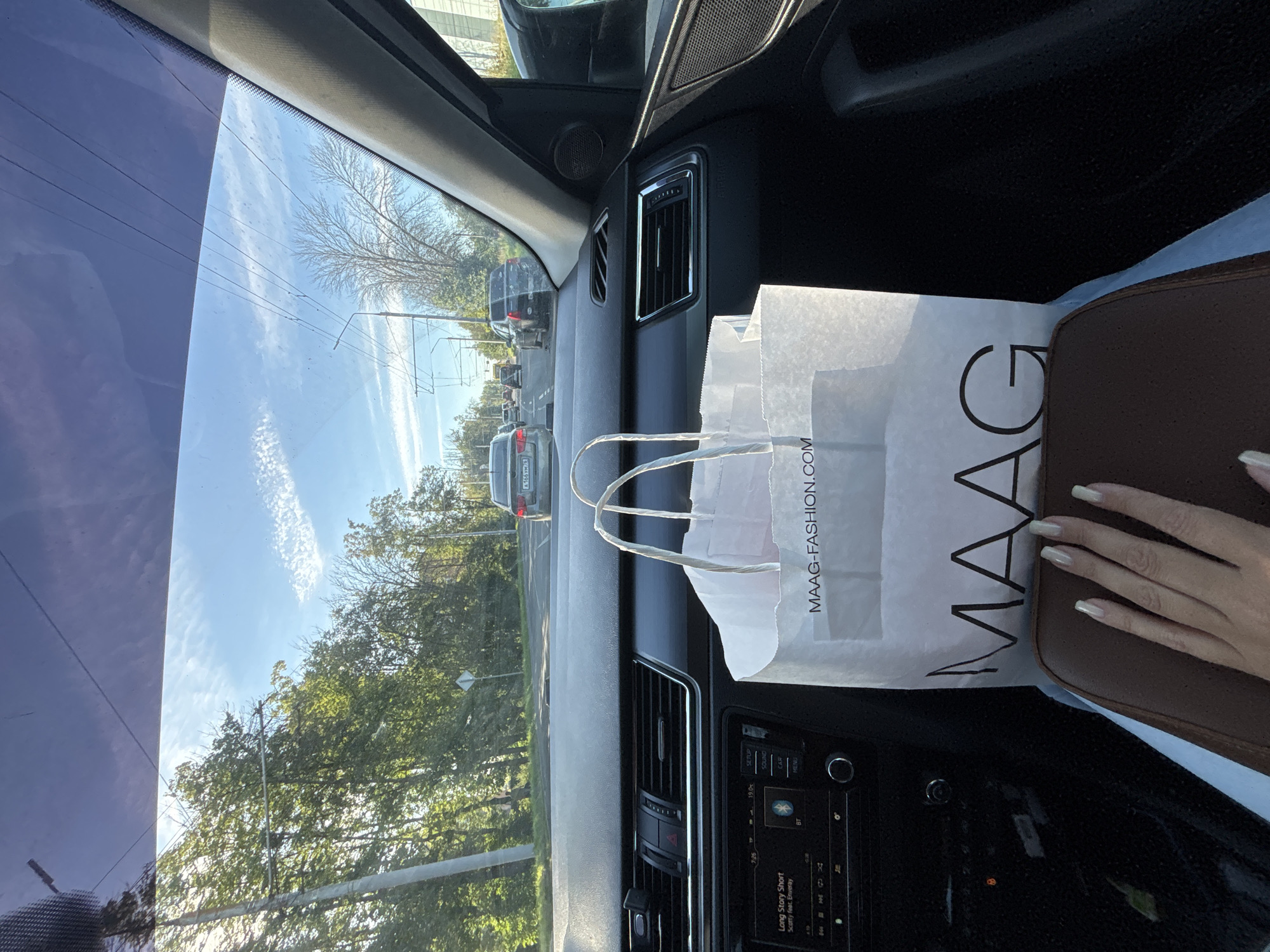 An exterior shot from inside a car captures a daytime view of a road in Zhukovo, Russia. The focus is on the dashboard and a shopping bag, with the road and surrounding trees visible through the windshield. The bag, made of white paper, displays the logo "MAAG" and the website "MAAG-FASHION.COM". A person's hand with light-colored fingernails rests on a brown surface in the foreground. The car appears to be in traffic, with other vehicles visible on the road ahead. The environment is characterized by a bright sky with scattered clouds and lush green trees lining the roadside.