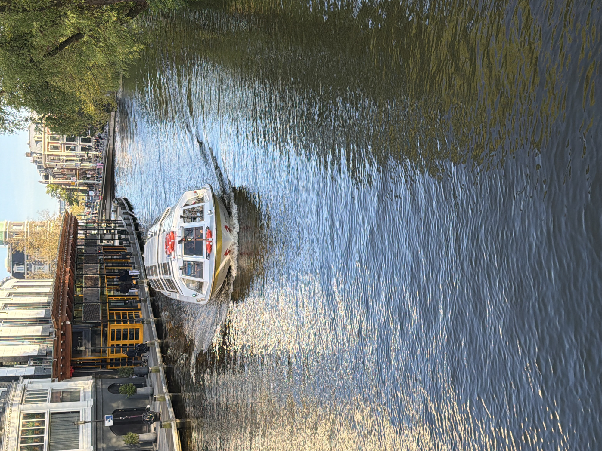 The image depicts a canal in Amsterdam, Netherlands. A white and gold tour boat is traveling along the canal, creating a wake in the water. On the left bank, there are buildings, including a restaurant with outdoor seating. People can be seen walking along the canal side. The water is reflecting the sunlight and buildings. Trees line the far bank.
