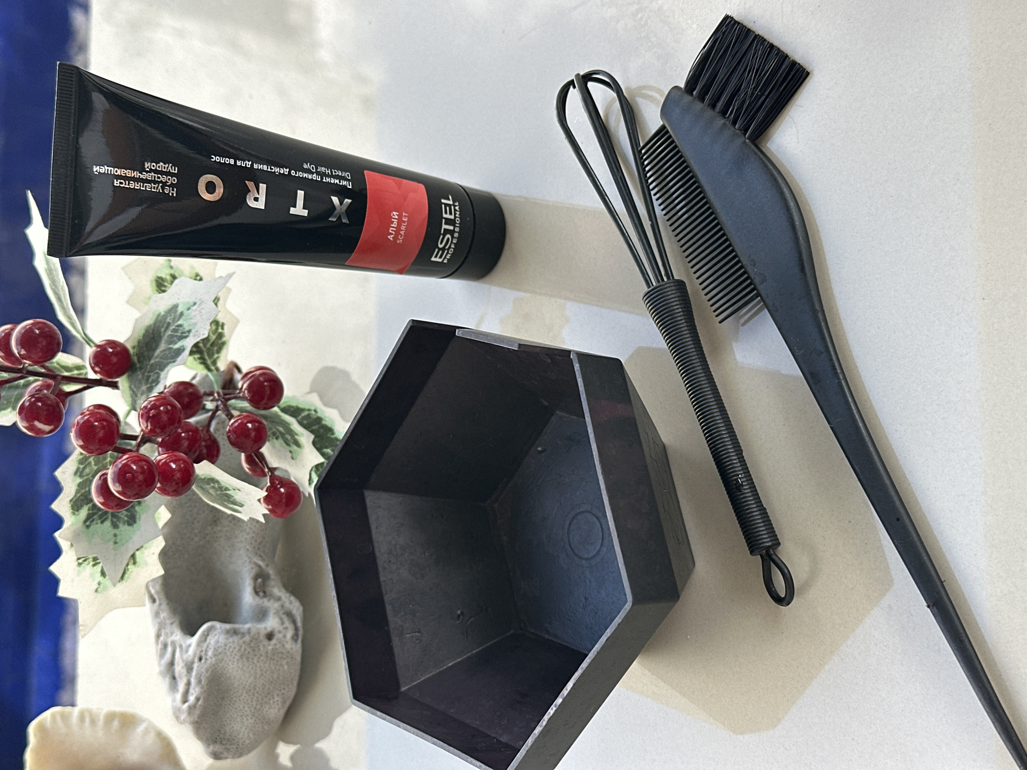 The image shows a collection of hair dyeing equipment laid out on a neutral surface. A black tube of "Estel Xtro" hair dye (scarlet) is positioned near the top left. Next to it is a decorative arrangement of artificial red berries and white/green leaves around a small, off-white vase or decorative item. To the right of the dye, a black, hexagonal mixing bowl sits prominently. A black whisk and a combination brush/comb (also black) are placed adjacent to the mixing bowl. The whisk is positioned at the right of the bowl, with the brush/comb on top of it. The background is a light beige or cream color.
Given the text on the hair dye product ("ESTEL PROFESSIONAL") and the presence of hair dyeing equipment, the scene likely relates to hair styling or cosmetic preparation, possibly in a salon or home setting. There is no evidence for the location in the city of North Kuta, Indonesia.