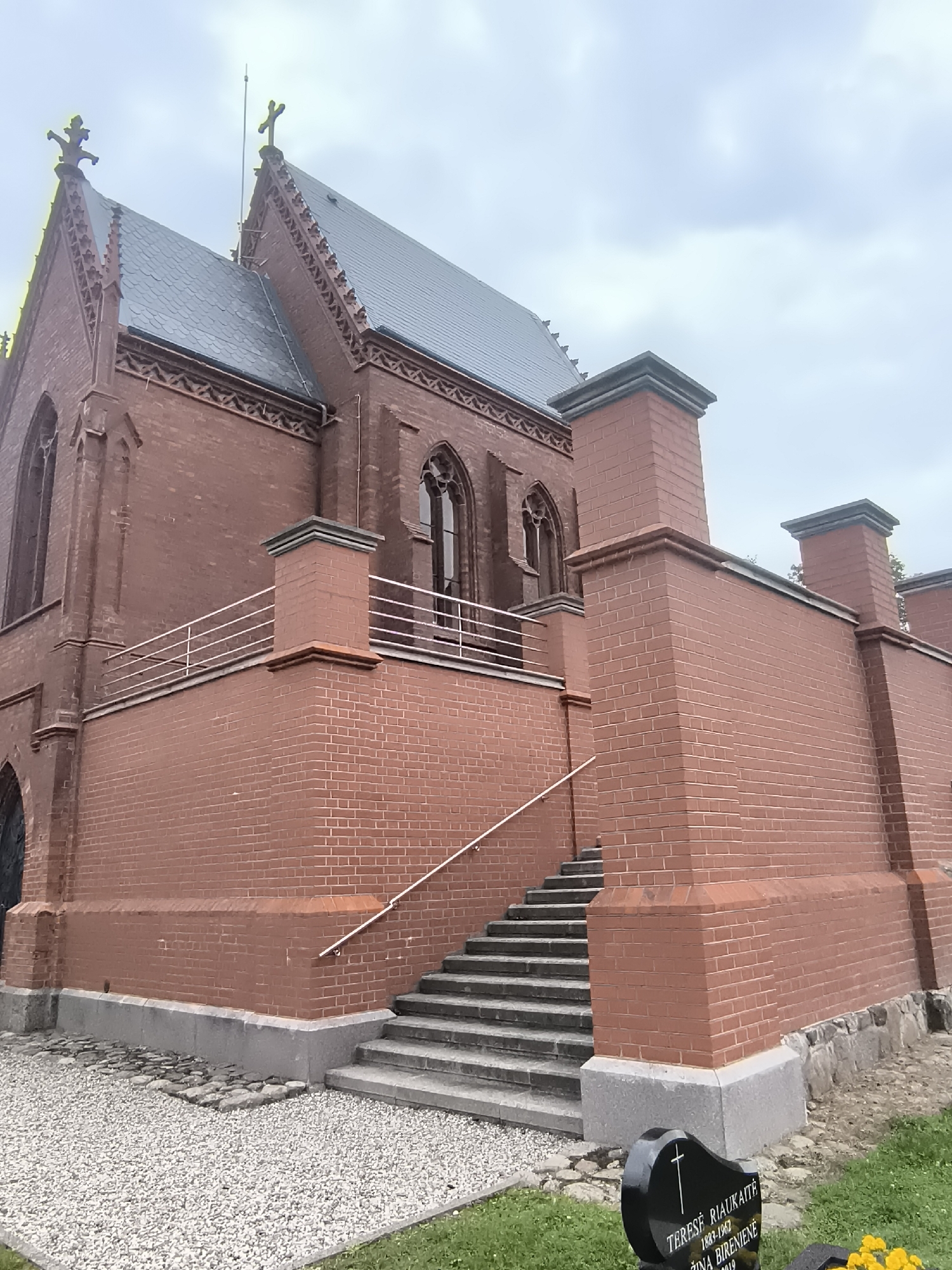 The image shows the side and rear of a brick building, likely a chapel or mausoleum, with a dark grey roof. The building features Gothic-style architectural elements such as arched windows with tracery and decorative brickwork along the roofline. A low wall with a metal railing runs along a raised section of the building, accessible by a set of outdoor stairs. The surrounding area is paved with gravel and some grass, and a black headstone with a cross and inscriptions is visible in the foreground. The sky is overcast. This structure is the Kretinga Catholic Cemetery Chapel in Kretinga, Lithuania.