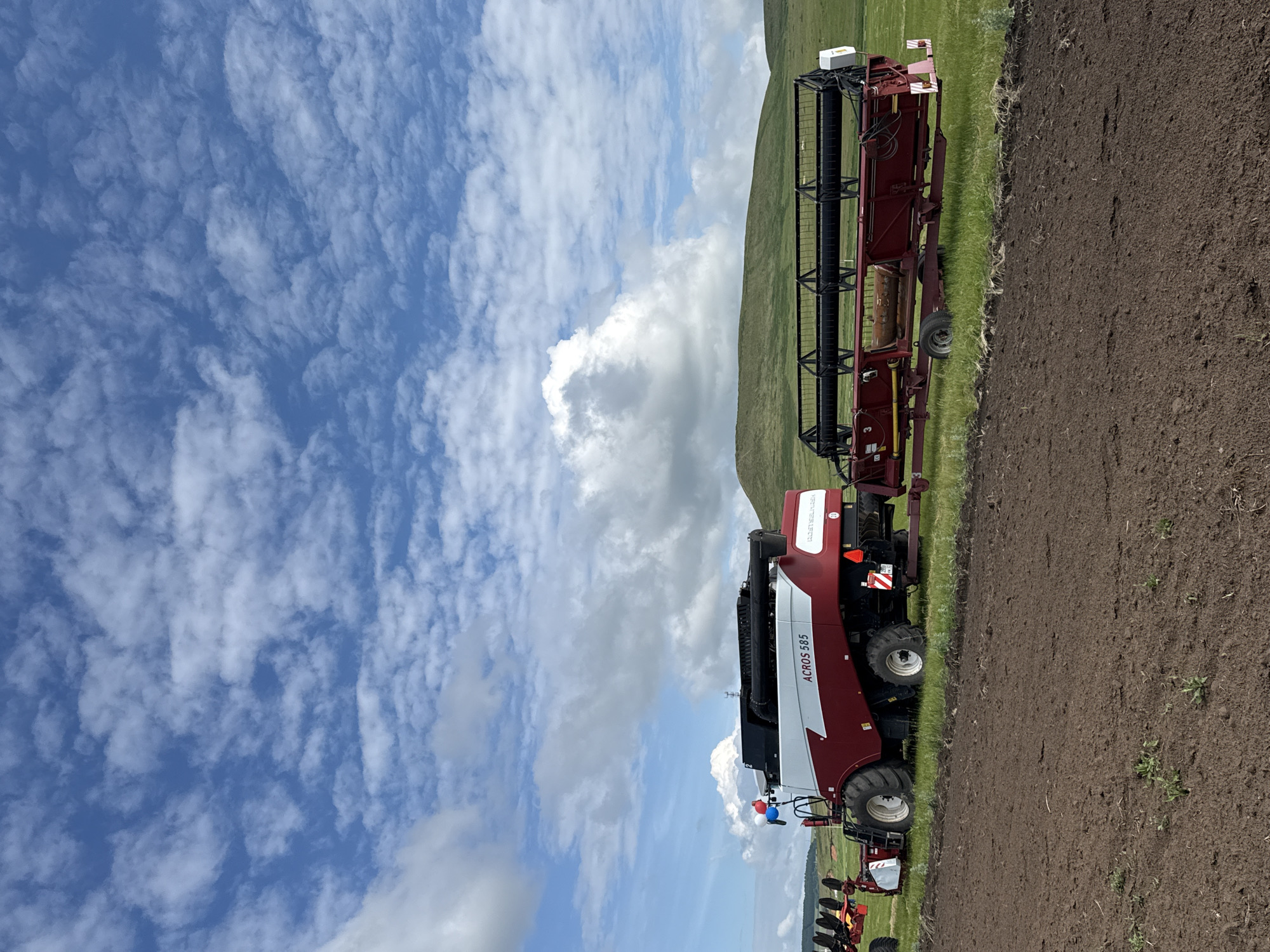 A combine harvester and its header attachment are parked on a field in Старое Субханкулово, Russia. The combine is red and white with "ACROS 585" written on its side. The header, also red, is attached to the front of the combine. The field is brown and appears to be recently tilled. In the background, there are green hills under a blue sky with scattered white clouds. The scene suggests agricultural activity.