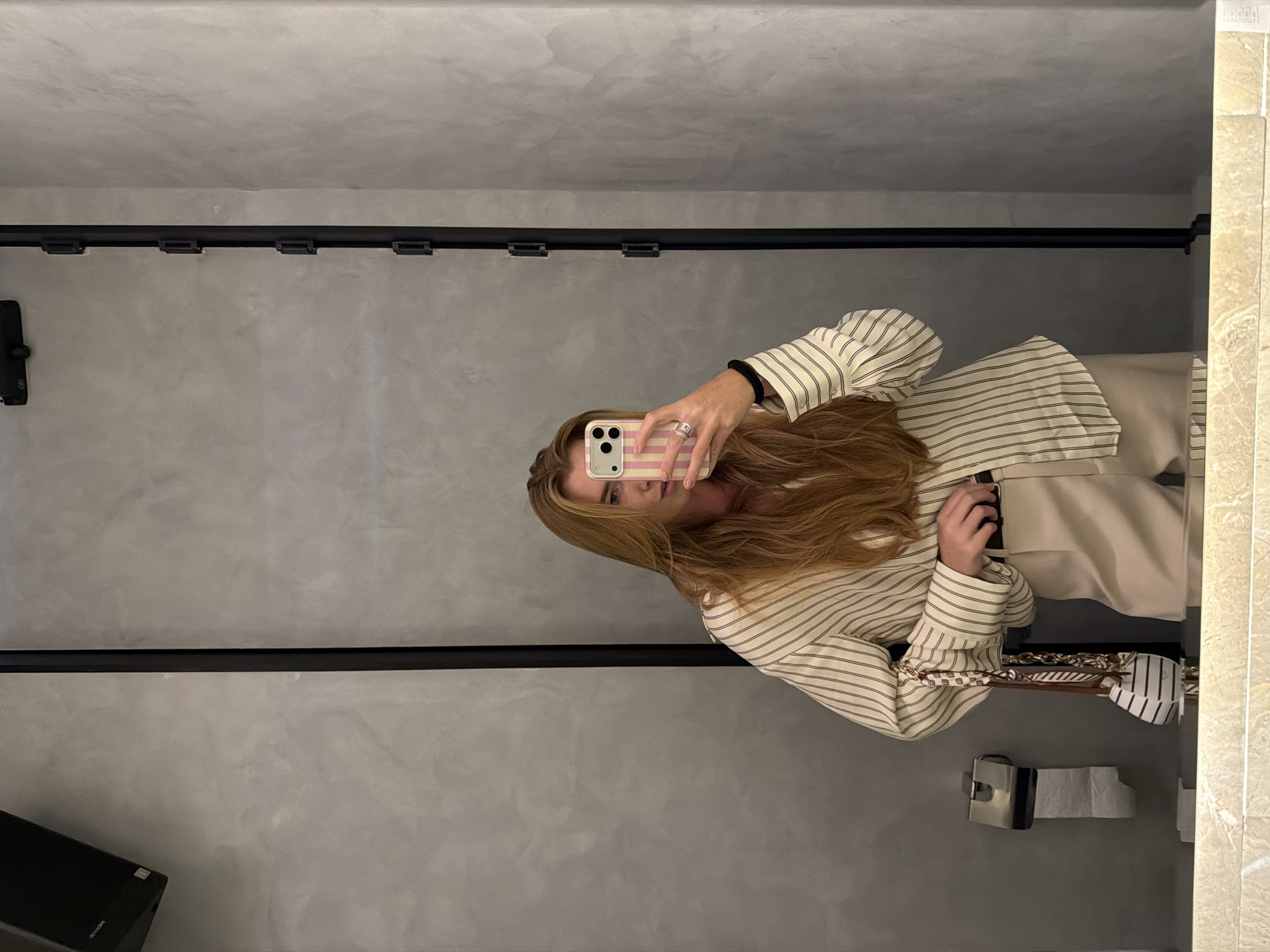 A person with long brown hair is taking a mirror selfie in a bathroom with grey walls. The person is wearing a striped shirt and beige pants. A black track lighting fixture runs along the top of the wall. A toilet paper dispenser is visible on the right side of the frame. The photo was likely taken in Bishkek City, Kyrgyzstan.