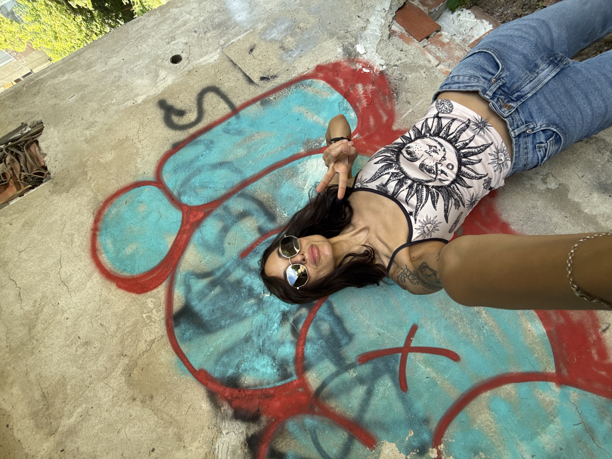 A woman is lying on a concrete surface with graffiti in an outdoor location. The graffiti is predominantly turquoise and red. The woman is wearing jeans and a tank top with a sun and moon design. She is also wearing sunglasses and has a tattoo on her arm. She is holding her hand up, giving a peace sign. In the background, there is greenery and what appears to be a building. The location is Pleven, Bulgaria.