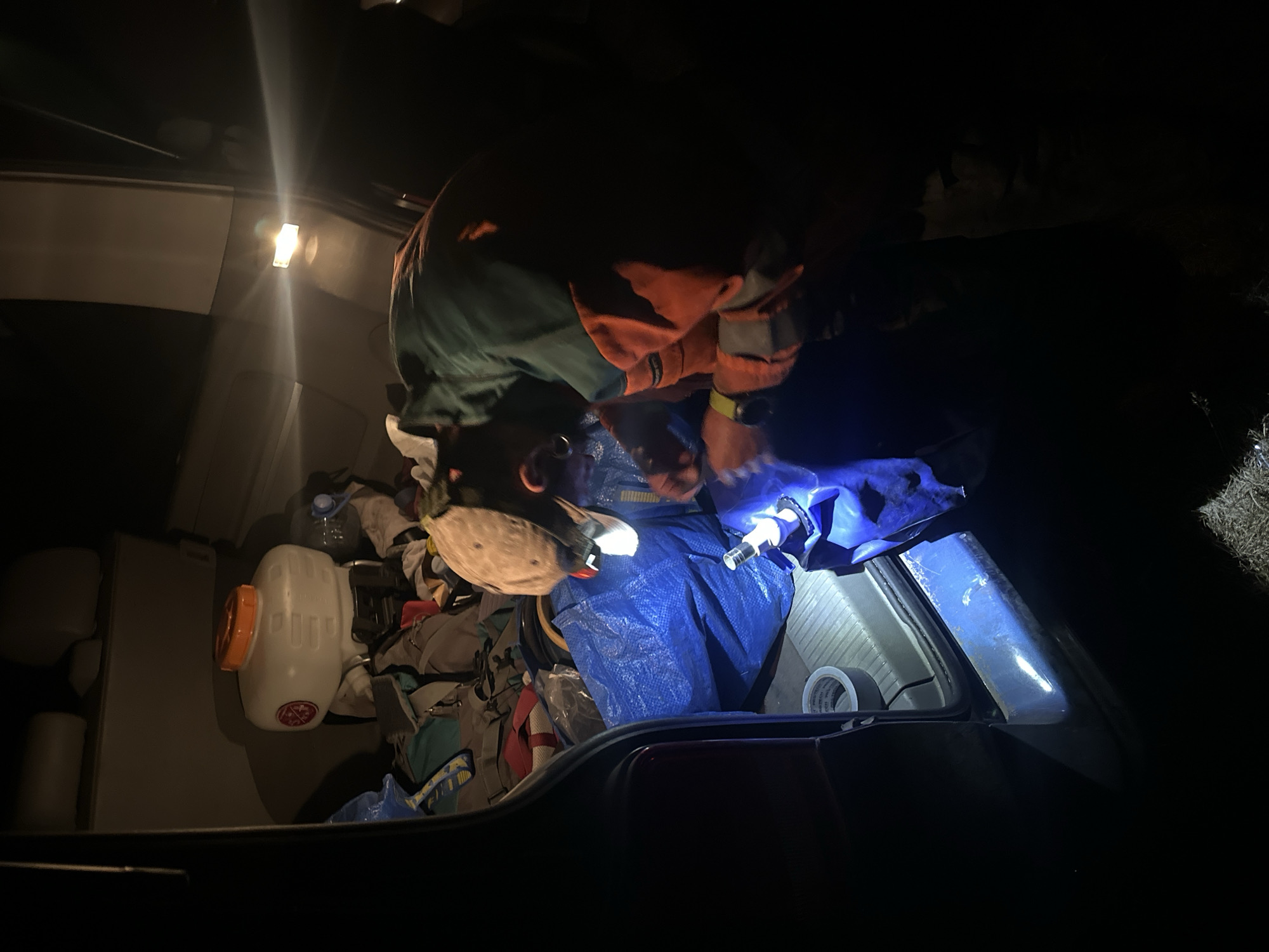In the cargo area of a vehicle, a person is seen wearing a green and orange jacket and a brown hat, tending to a blue tarp. The vehicle is loaded with various items, including a white plastic tank with an orange lid, several bags, and what appears to be duct tape on the floor. The scene is dimly lit, with a bright light source illuminating the area where the person is working. The context suggests a scene in Bijele Poljane, Montenegro, likely involving some form of outdoor activity or transport of goods.