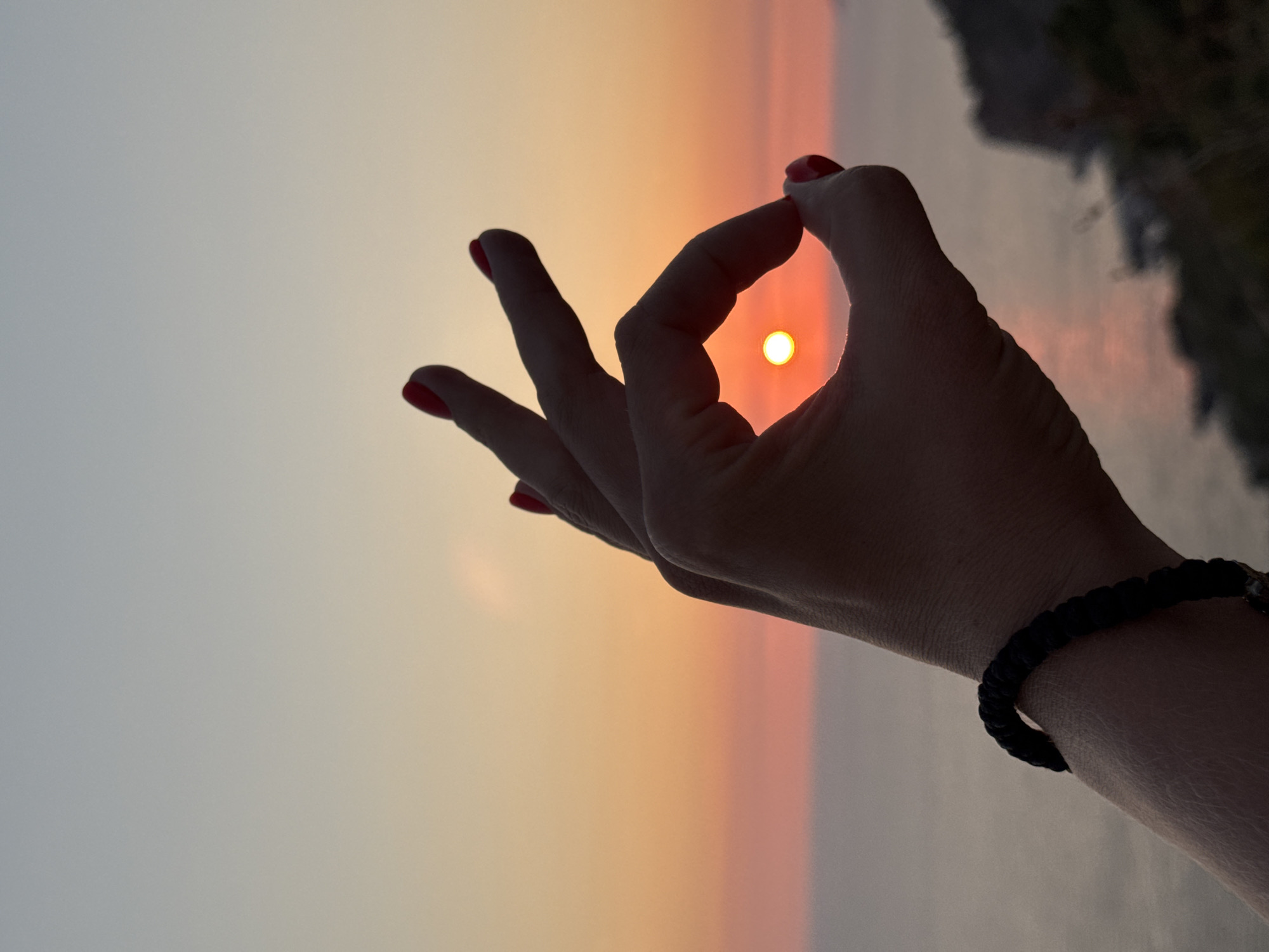A silhouette of a hand with bright red nail polish forms a circle with its thumb and index finger, framing a small, glowing sun setting over the Adriatic Sea. The sun is a vibrant orange-yellow orb, appearing as if it is being held within the hand. The sky transitions from a pale blue at the top to soft hues of pink and orange near the horizon, indicating the time of sunset. The hand is adorned with a dark braided bracelet on the wrist. In the background, the calm sea stretches out, meeting the horizon line. On the right side of the image, the blurred outline of a rocky coastline or cliff face is visible, with some dark vegetation. The overall scene suggests a scenic overlook, likely in a coastal area, at dusk. The provided information mentions Donji Katun, Montenegro, which is inland and not directly on the coast, making the location description potentially inaccurate for the visual scene.