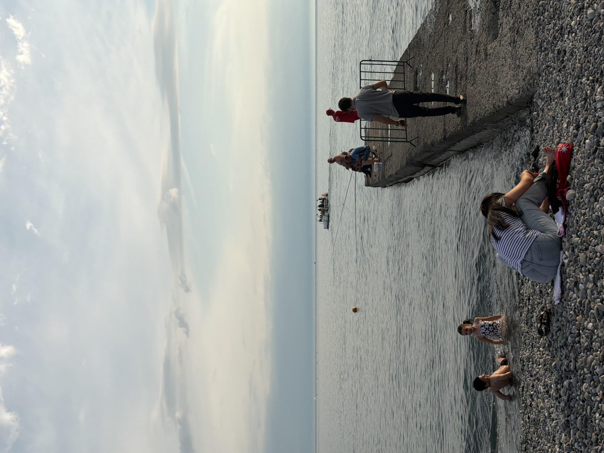 The image depicts a serene coastal scene in Sochi, Russia, with the Black Sea stretching towards the horizon under a pale, cloudy sky. In the foreground, on a pebble beach, a couple sits facing away from the camera, with the woman resting her head on the man's shoulder. To their left, two children play in the shallow water. Further out in the sea, a small boat carrying several people is visible, along with a lone orange buoy floating on the water. A concrete pier extends into the sea, where a group of people are fishing. A man in a grey shirt walks away from the camera on the pier, towards the sea. The overall atmosphere is one of relaxed leisure and summer tranquility.
