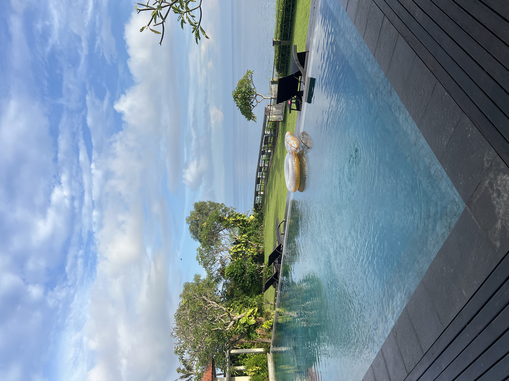 An outdoor scene captures a luxurious infinity pool area under a bright, partly cloudy sky, characteristic of a tropical location like Kuta Selatan, Indonesia. The clear blue water of the swimming pool dominates the right and central portions of the image, with dark wooden decking forming its immediate edge on the right side. Two golden, decorative inflatable rings float leisurely in the pool.
Beyond the pool, a vibrant green lawn stretches out, dotted with lush tropical vegetation, including several trees with dense foliage and what appears to be a distinctive frangipani tree. Two dark, modern sun loungers are positioned on the grass, facing towards the distant horizon. A railing or fence separates the lawn from a vast, calm body of water, likely the ocean, which extends to the horizon under the bright sky. The sky itself is a vivid blue, adorned with scattered white, fluffy clouds, suggesting clear and pleasant weather conditions, likely during late morning or early afternoon. No people or visible text are present in the image, conveying a sense of serene tranquility and an inviting, private resort-like atmosphere.