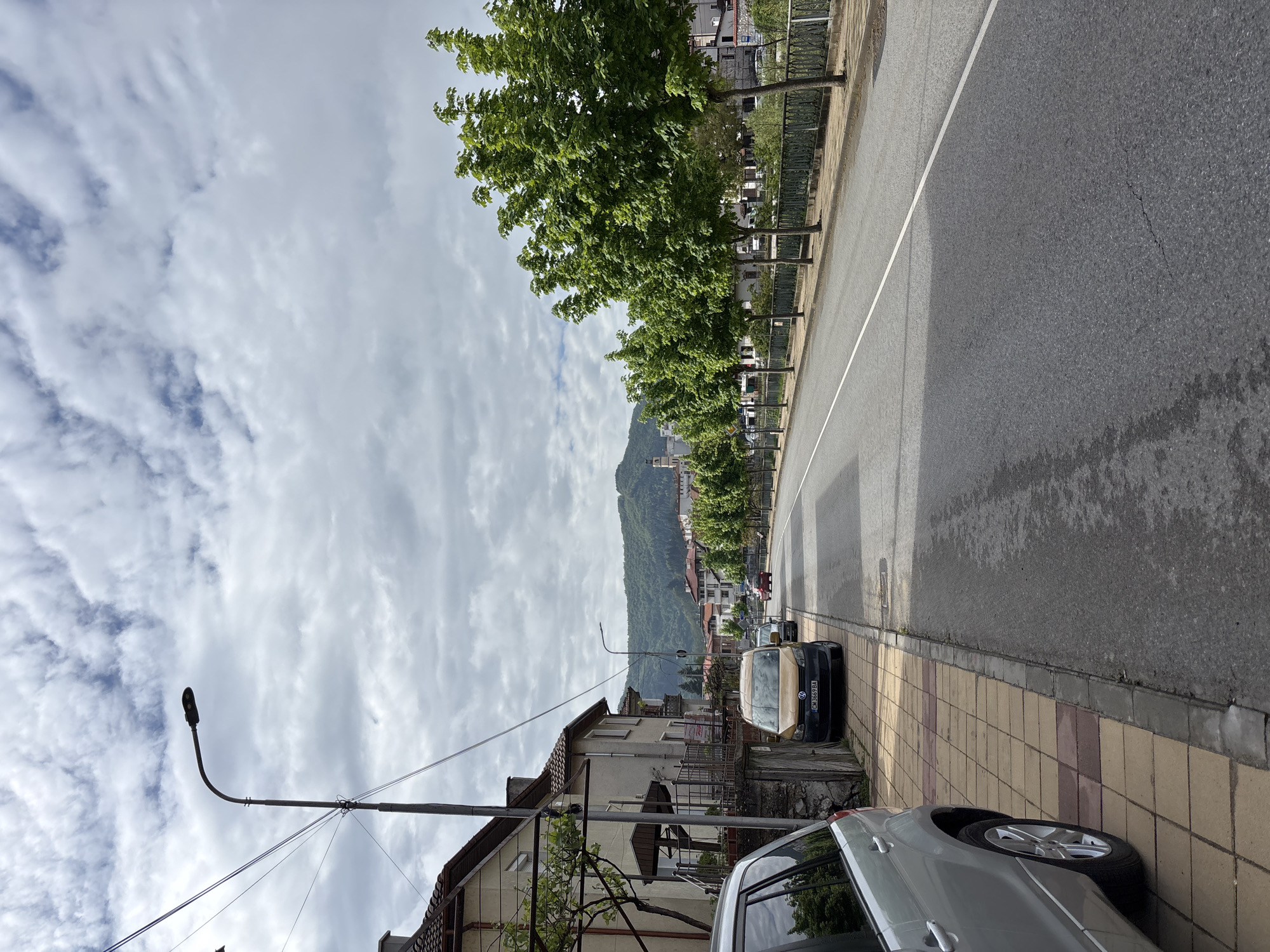 The image shows a street in Zlatograd, Bulgaria, with a view toward a mountain range in the distance. The sky is overcast with patterned clouds. The street is paved and has a white line marking the lane separation. A row of trees lines one side of the street, adjacent to a fence. Cars are parked along the sidewalk. A silver car is partially visible in the foreground, and a black car is parked further down the street, near a light pole. The sidewalk is paved with light-colored tiles. A building is visible behind the light pole. Other buildings can be seen in the distance along the base of the mountain.