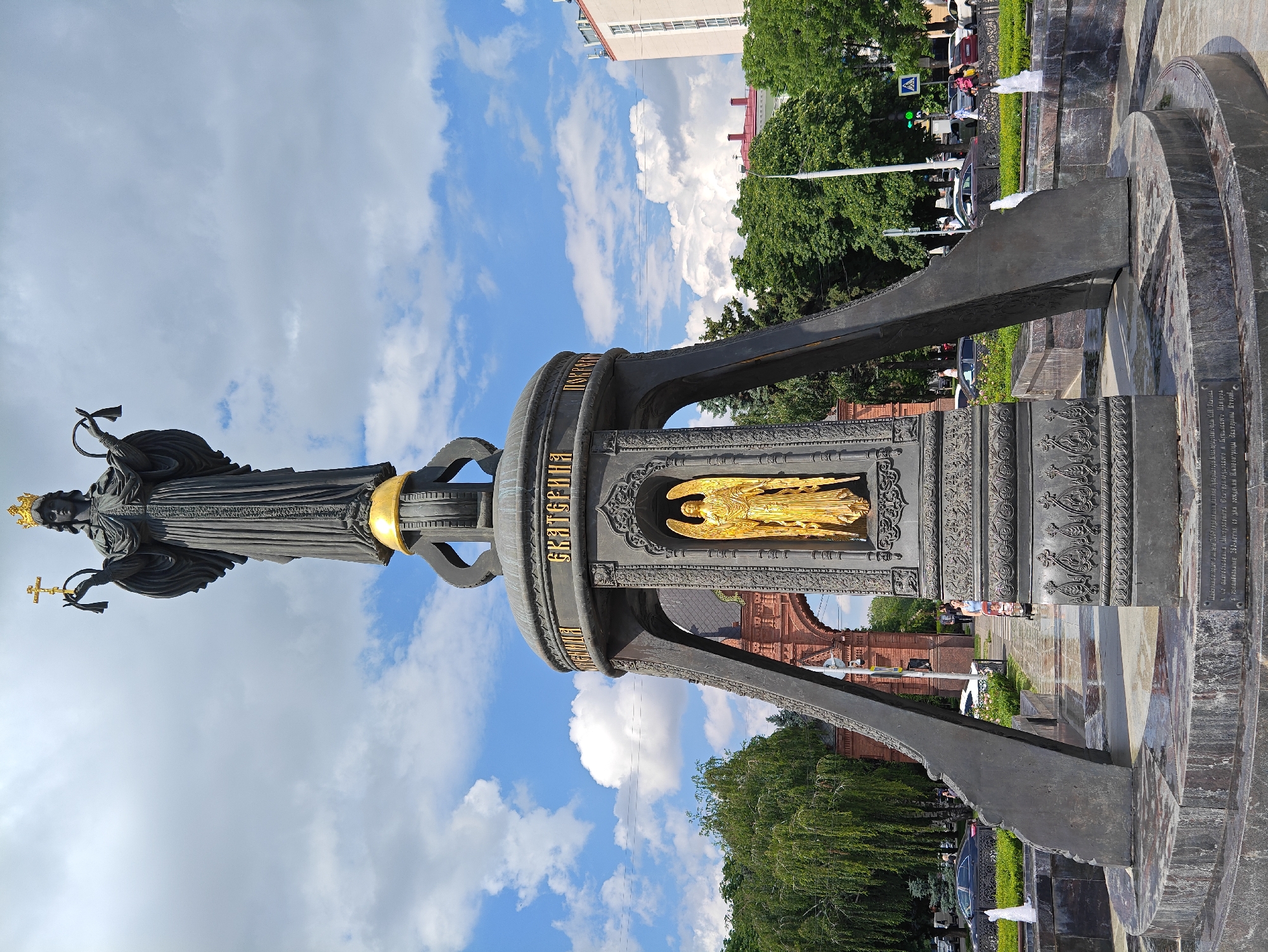 The image displays a monument in Krasnodar, Russia, dedicated to Empress Catherine the Great. The monument is a large, dark metal sculpture featuring a statue of the empress at the top, holding a cross and an orb. Below her, a golden angel is visible within a decorative niche. The base of the monument is surrounded by a fountain with water jets. In the background, there is a red brick archway, trees, and buildings, suggesting an urban setting. Several people are walking around the monument and in the background, and cars are visible on the street. The sky is blue with white clouds, indicating daytime. The inscription on the monument's base is in Russian.