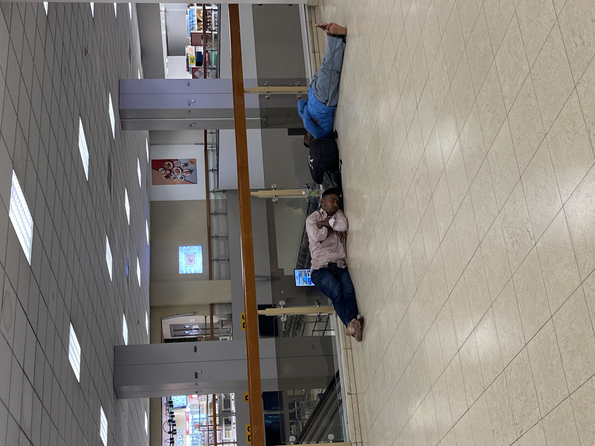 In a well-lit indoor space with a tiled floor, two men are resting on the floor. The man in the foreground is lying on his side, asleep, with his head resting on his arm. He is wearing a light-colored, patterned shirt and blue jeans. The second man, further back and also asleep, is lying on his back with his legs outstretched. He is wearing a blue jacket over a dark shirt and grey pants. A black backpack is placed next to him. The setting appears to be an airport lounge or a similar public waiting area, suggested by the tiled flooring, the overhead lights, and the presence of what might be shops or concessions in the background, visible through glass partitions. The overall impression is one of weary travelers taking a moment to rest. There is no visible text in the image.