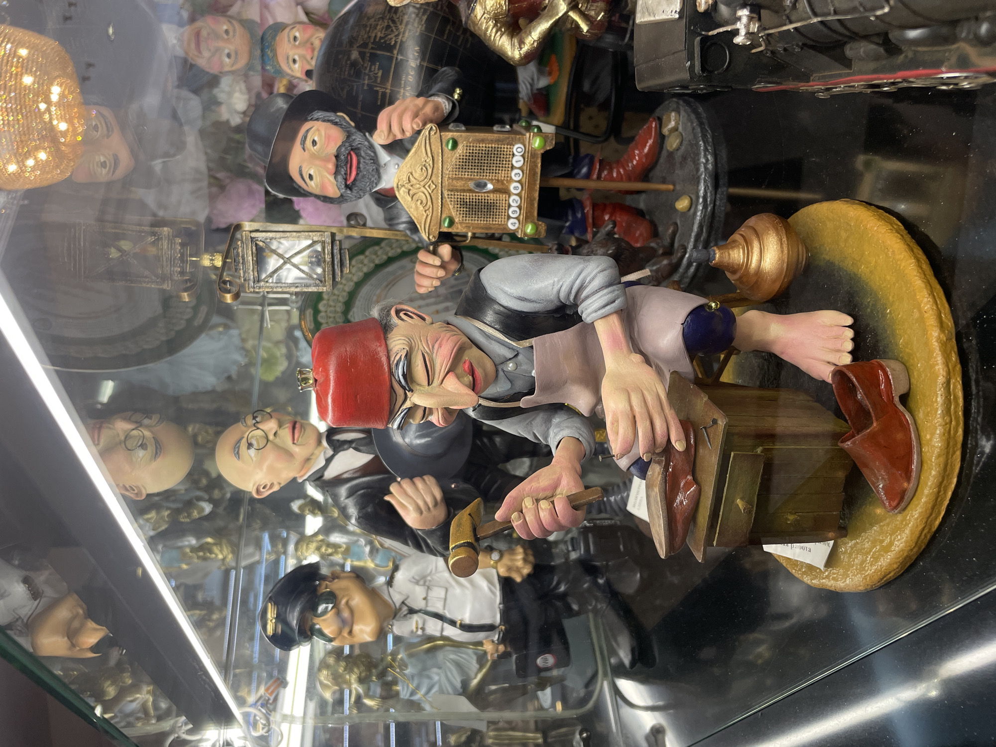 The image shows a display case with several figurines. In the foreground, a figurine of a cobbler, wearing a red fez, a light-colored apron over a grey shirt, and blue pants, is depicted hammering a nail into a shoe that rests on a wooden workbench. The cobbler has bare feet. To the left of the cobbler, a figurine dressed as a pilot with a black cap and sunglasses stands holding a briefcase. Behind them, a figurine in a black suit and tie stands holding a small, ornate wooden music box. A black train engine figurine is visible on the right side of the display. The figurines appear to be made of resin or similar material and are displayed on a reflective surface. The background of the display case shows other figurines and decorative elements, as well as reflections of lights and what appear to be other display cases.
The image is a photograph of a display of souvenir figurines. The specific location within the city of Kazan, Russia, cannot be determined from the image alone.