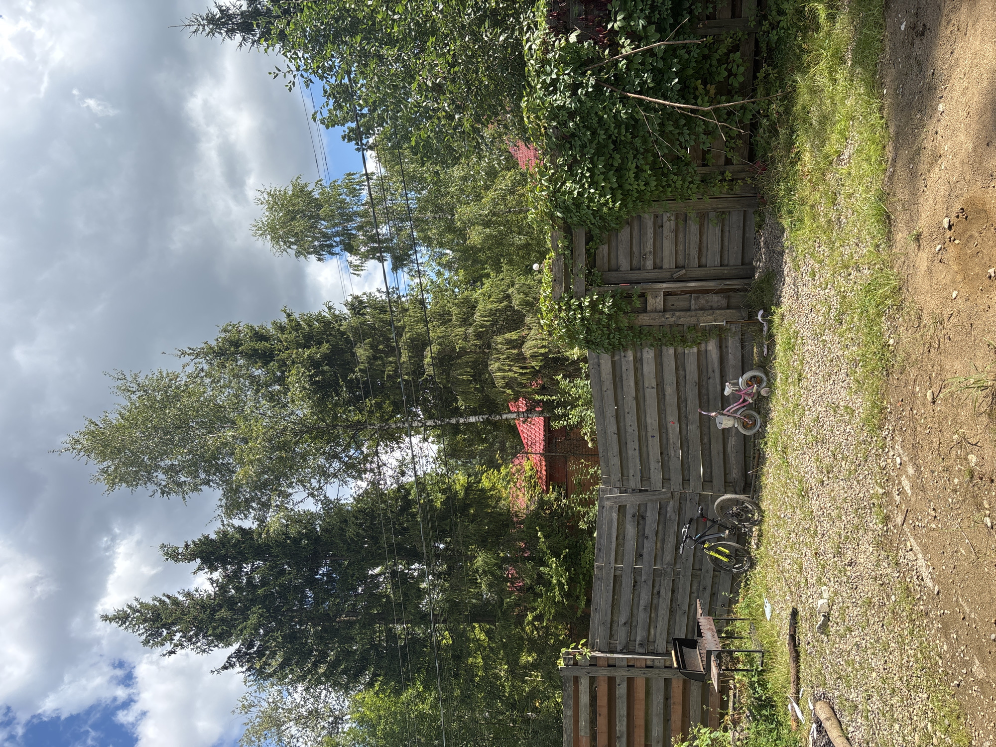 The image depicts a rustic, outdoor scene, likely a backyard or yard area. Dominating the background are tall evergreen trees and deciduous trees, with a section of red-tiled roof visible through the foliage, suggesting the presence of a building or house. A wooden fence, constructed from horizontal planks, runs across the midground, with a gate on the right side.
In the foreground, there is a gravelly and dirt ground surface, with patches of grass growing. To the left, a metal barbecue grill is set up. Two bicycles are parked near the fence: a larger, darker colored bicycle with yellow accents and a smaller pink bicycle, presumably for a child. To the far left, a pile of logs suggests a fire pit. The sky above is partly cloudy, with a mix of bright blue patches and gray clouds.
The location is identified as Granat, Russia. The scene appears to be a residential property, possibly in a rural or semi-rural setting, given the natural elements and the style of the fence and building. There are no people visible in the image. The overall impression is one of a quiet, somewhat unkempt outdoor space.
