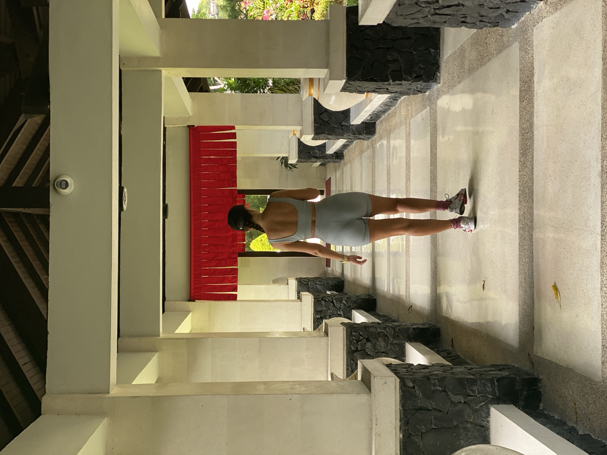 A woman is standing in a colonnade in Kuta Selatan, Indonesia. She is facing away from the viewer and is wearing a light blue athletic top and shorts and sneakers. The colonnade has white columns with black stone accents. A red curtain is hanging in the background. The floor is a light-colored stone with dark stone accents. A security camera is visible on one of the columns. There is foliage visible in the background.