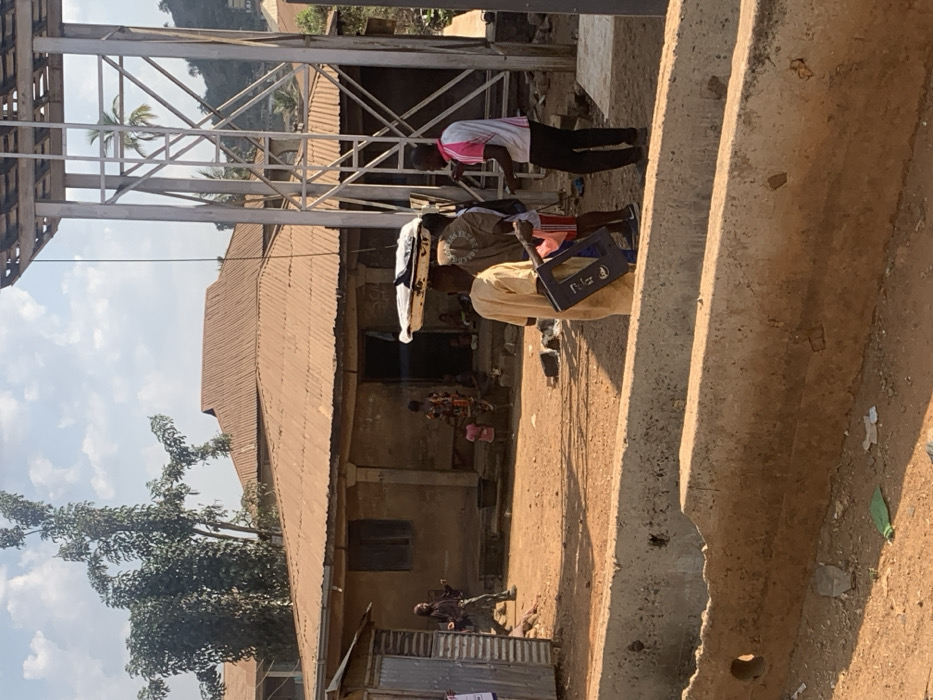 The image depicts an outdoor scene in Egbedi, Nigeria, under a bright, partly cloudy sky. The ground is composed of reddish-brown dirt.
In the mid-ground, a single-story building with light-colored, possibly stucco or mud walls and a rust-colored corrugated metal roof dominates the left and center. It features multiple windows, some with visible shutters, and a central doorway where several individuals are gathered. One woman in a colorful patterned dress and two children in pink clothing are discernible near this entrance. To the far left of this building, a smaller, corrugated metal shack is visible.
To the right of the main building, a tall, grey metal lattice tower stands with a wooden platform at its apex. A thin cable or wire extends downwards from the tower's structure. In the foreground, two parallel, weathered concrete barriers or channels run horizontally across the lower portion of the frame. Scattered debris, including green plastic and white paper, is visible on the dirt ground between and around these barriers.
Several individuals are present. In the right-center mid-ground, a man in a light-colored, long traditional garment (possibly an agbada) holds a blue plastic crate. Adjacent to him, another man balances a white tray containing dark objects on his head, wearing a dark shirt and shorts. A third man in a white and pink polo shirt and dark pants stands facing these two, appearing to interact with them. Further left, in front of the main building, a person in dark, possibly camouflage-patterned clothing with a facial covering walks towards the left, away from the building. In the background, beyond the main building, more structures with similar corrugated roofs are visible, along with a hillside featuring sparse vegetation including palm trees. A large tree with green foliage stands to the left of the main building.