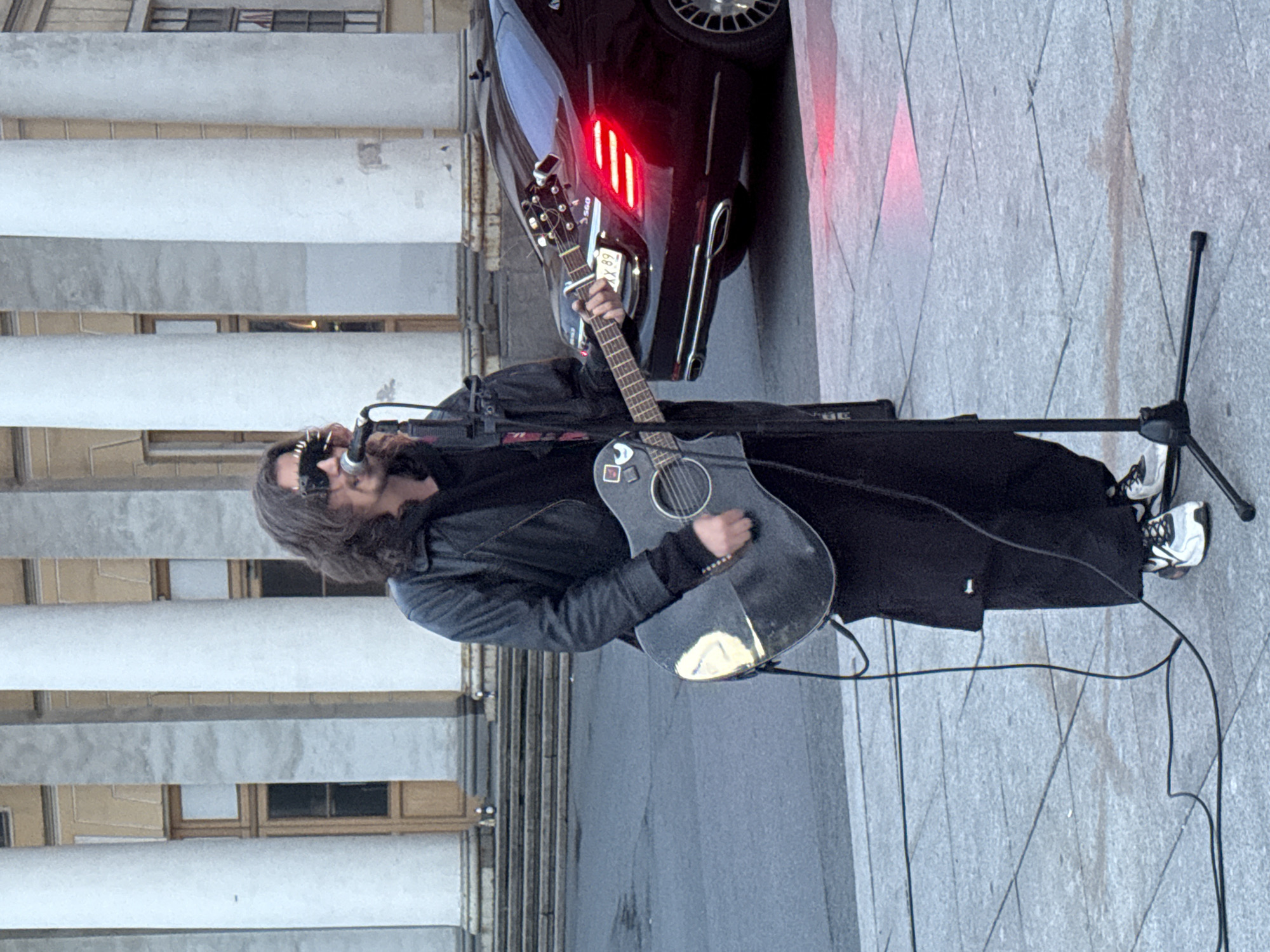 A man is playing an acoustic guitar and singing into a microphone on a paved outdoor plaza in front of a building with large white columns. The man has long, dark hair and is wearing sunglasses adorned with spikes. He is dressed in a black leather jacket and black pants. The guitar is dark gray with some stickers on it. A microphone stand is positioned in front of him. In the background, a black luxury car with its brake lights on is parked. The architecture suggests a historic European city, and the car's design, particularly the taillights and grille, is consistent with a modern Mercedes-Benz S-Class. The overall scene indicates a public performance. The location is likely Saint Petersburg, Russia, given the architectural style and the presence of the car, which is common in the city.