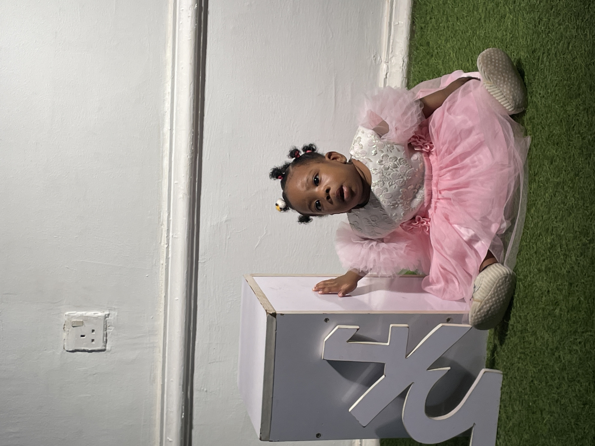 A young child, likely a girl, is captured in an indoor setting, possibly a studio, in Barakin Akawo, Nigeria. The image is rotated 90 degrees clockwise, showing the child positioned next to and on a white/light grey box or platform. She looks directly at the camera with a neutral to slightly curious expression, her head tilted.
The child has dark skin and her hair is styled in several small braids or knots, adorned with colorful ties. She is dressed in a festive outfit consisting of a white top with intricate, possibly embroidered or sequined, patterns, and large ruffled sleeves. A voluminous, fluffy pink skirt or dress, appearing to be made of tulle, completes her attire. Light-colored, possibly white or cream, shoes are visible on her feet. Her right hand rests gently on the top surface of the box.
On the side of the box, large, light grey letters with a white border are partially visible, appearing to spell out "SKY". The background features a textured white wall with decorative vertical molding or trim on the left. An electrical outlet, consistent with a Type G socket commonly found in Nigeria, is visible on the wall. To the right, a vibrant green artificial turf or carpet covers the floor, adding a contrasting texture to the scene. The lighting suggests an indoor environment, likely lit by artificial light.