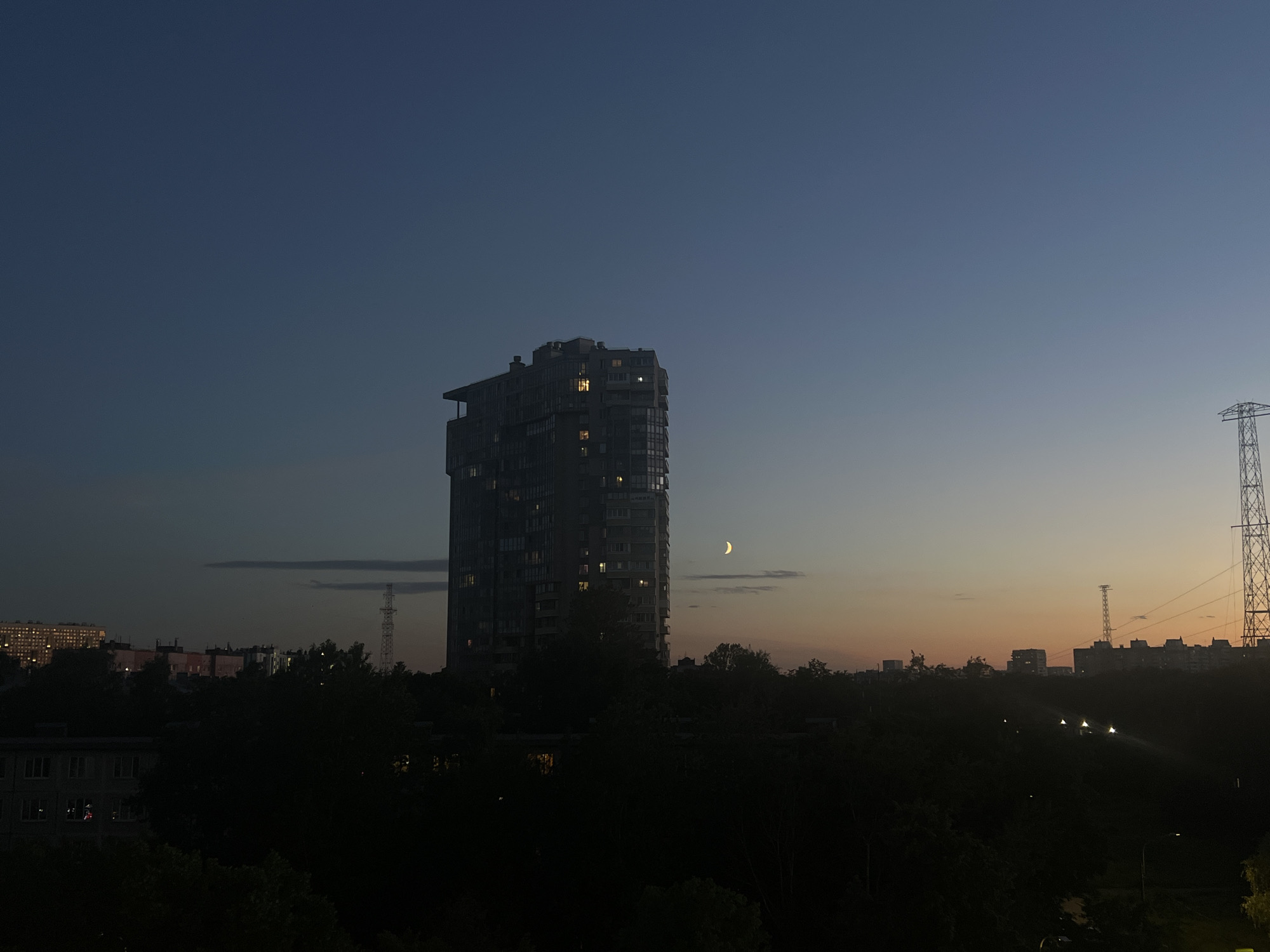 This image depicts a dusky cityscape in Sinyavino, Russia. In the foreground, dense, dark foliage obscures much of the lower portion of the scene. Above the trees, a tall, modern apartment building dominates the mid-ground, its windows illuminated with a warm yellow light. To the right of the apartment building, a slender crescent moon hangs in the twilight sky. Further in the distance, a series of lower buildings and structures are visible, some with illuminated windows. A tall, lattice-like power pylon stands prominently on the right side of the frame, its silhouette stark against the gradient of the sky, which transitions from deep blue at the top to a soft orange and yellow near the horizon. A faint, horizontal cloud streak is present in the upper left quadrant. The overall atmosphere is calm and serene, suggesting the transition from day to night in a residential urban area. There are no people or discernible actions in the image.