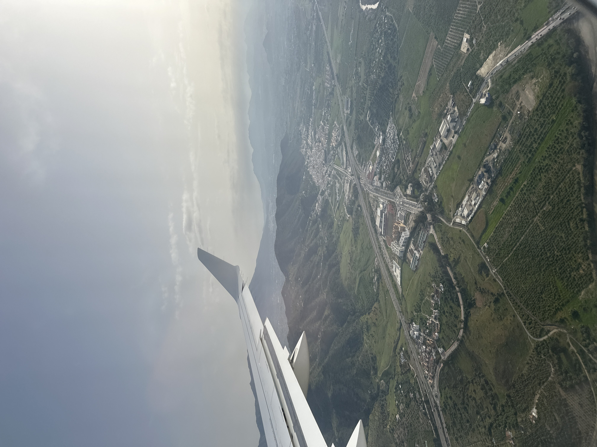 Aerial view of Almogía, Spain, from an airplane. The airplane's wing is visible in the lower-left corner. In the background are mountains shrouded in haze. The landscape includes green fields, clusters of buildings, roads, and highways.