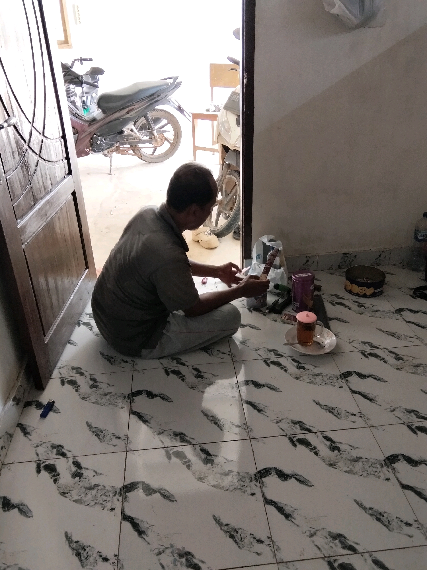 A man is sitting on the floor in front of a partially open wooden door, in what appears to be a residential area in Perambahan, Indonesia. The floor is tiled with a white and black marbled pattern. To his left, a motorcycle is parked just outside the doorway. In front of the man are several items, including a cup of tea on a saucer, a tin of cookies, a plastic bag, and what seems to be a pack of cigarettes. He is holding a cigarette in his right hand. The lighting suggests it is daytime.